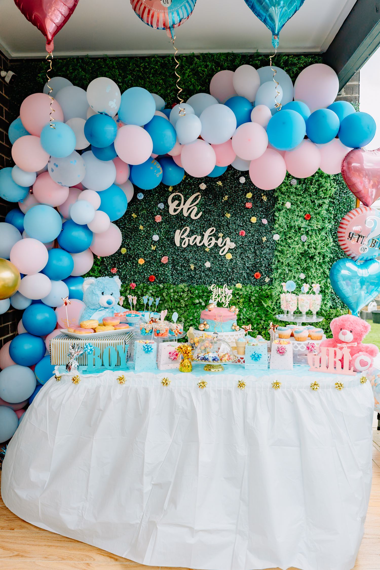 Pink and blue balloon arch decorates a gender reveal party dessert table with white tablecloth and greenery backdrop.