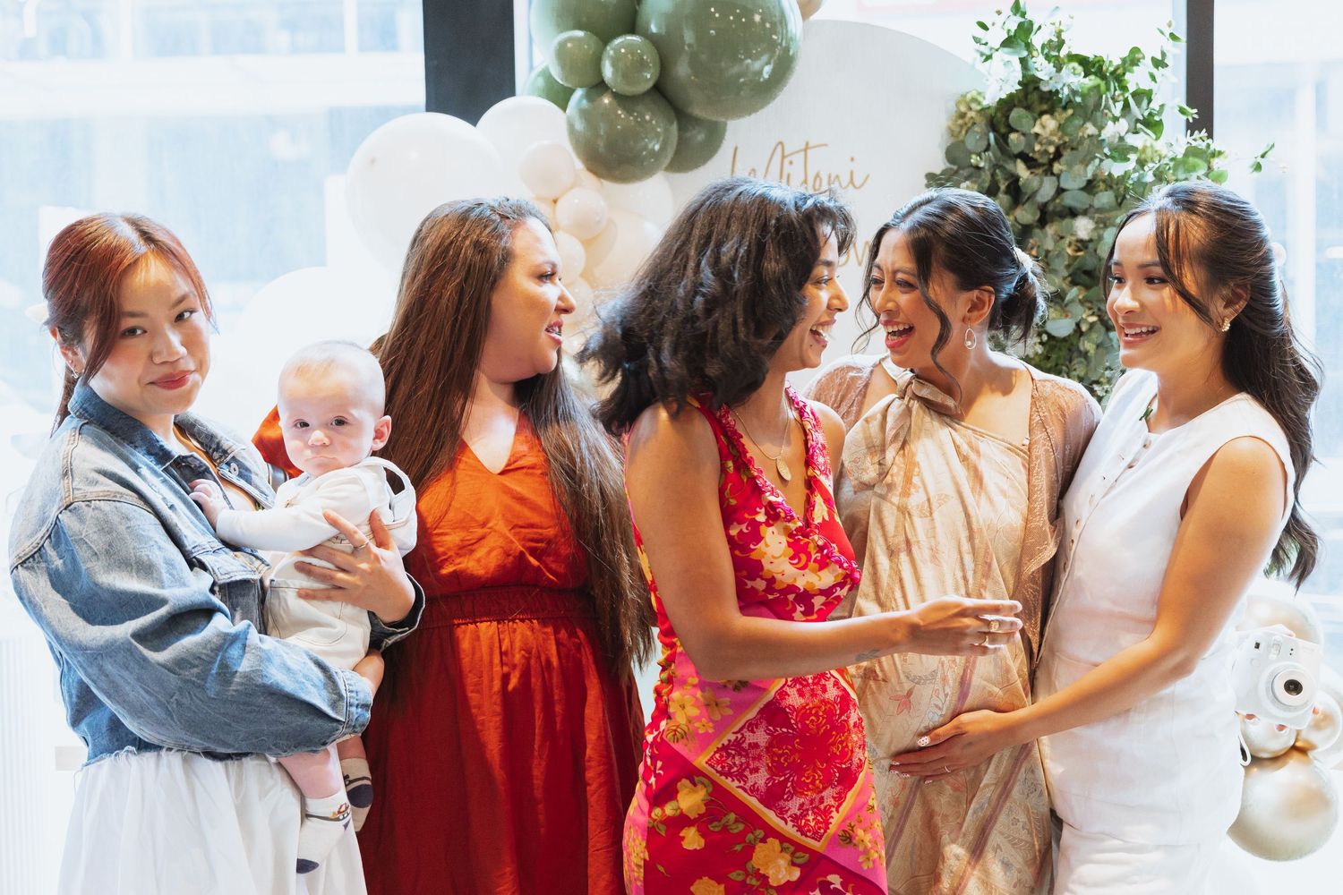 A group of friends in colorful dresses gather together at a celebration with a baby and decorative balloons.