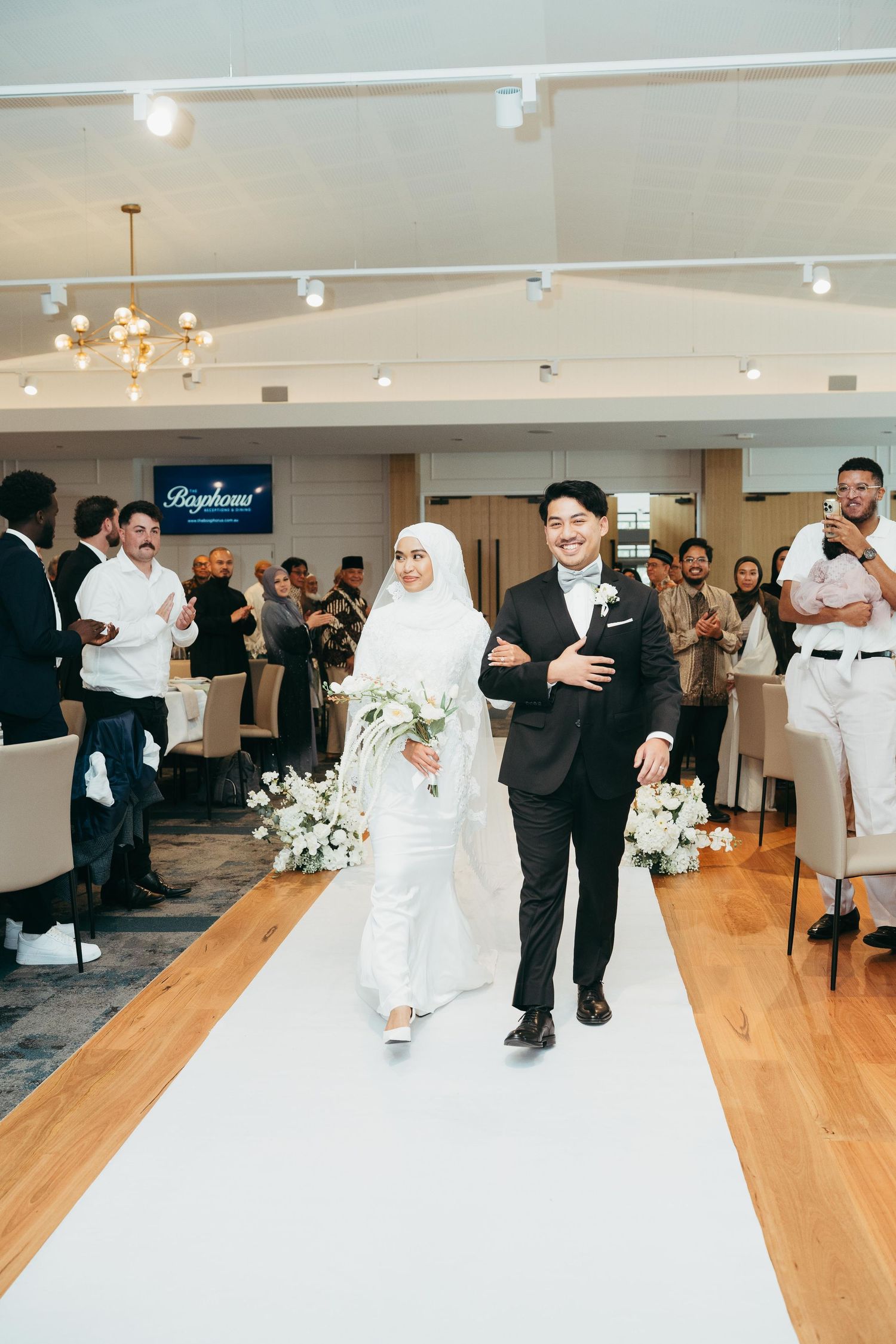Muslim bride in white hijab and groom in black tuxedo walk down the aisle at reception.