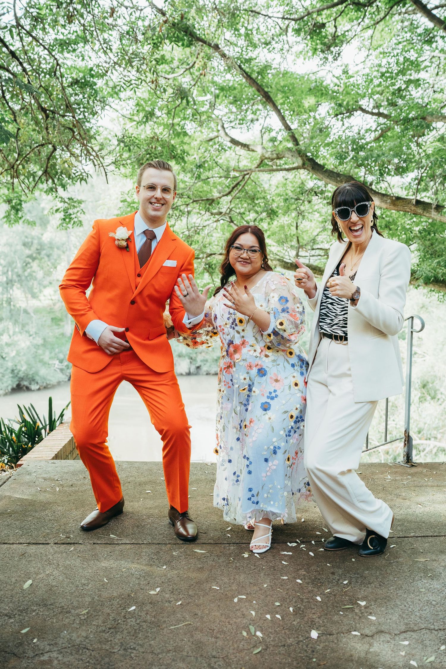 Groom in orange suit poses playfully with bride in floral dress and guest in white suit outdoors.