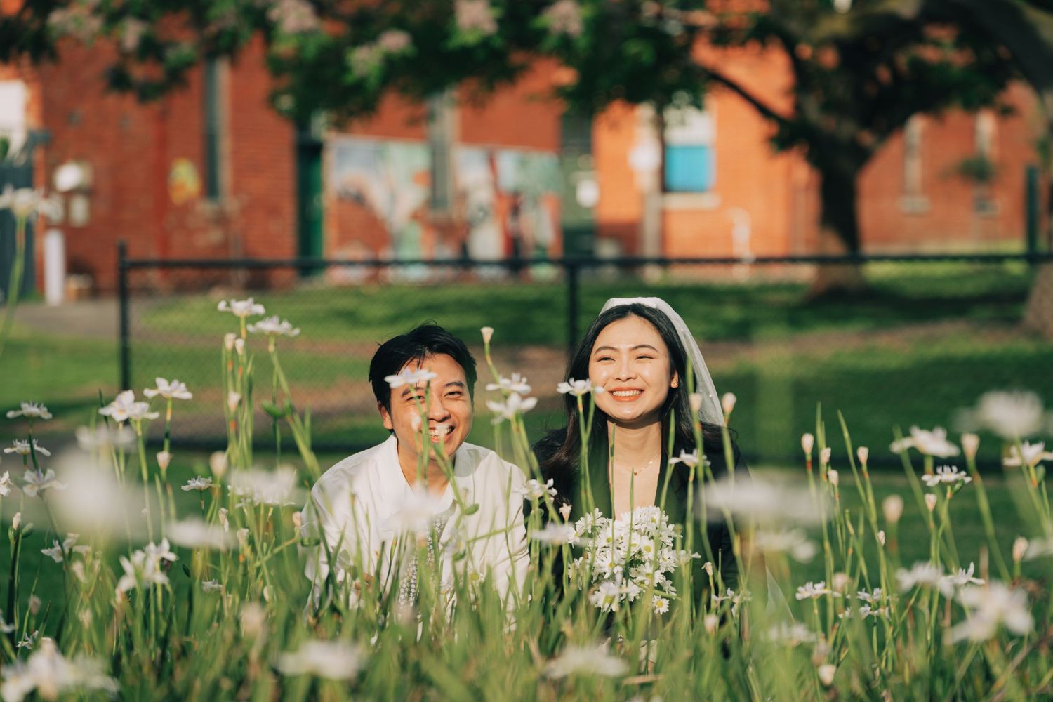 Couple smiling among white wildflowers in a lush green field with a red brick building in the background.