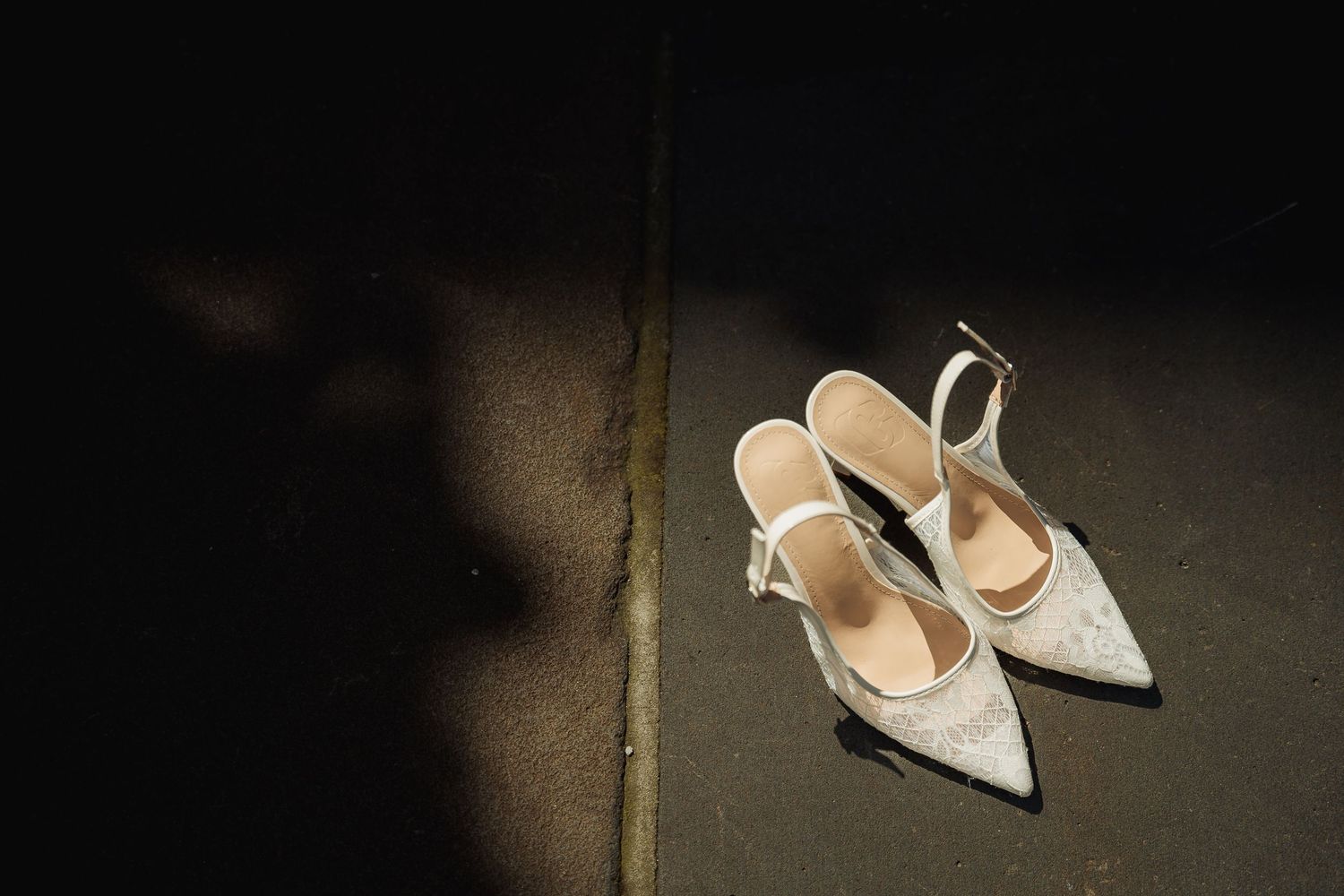 Elegant white bridal heels with pointed toes and ankle straps resting on dark floor in dramatic lighting.