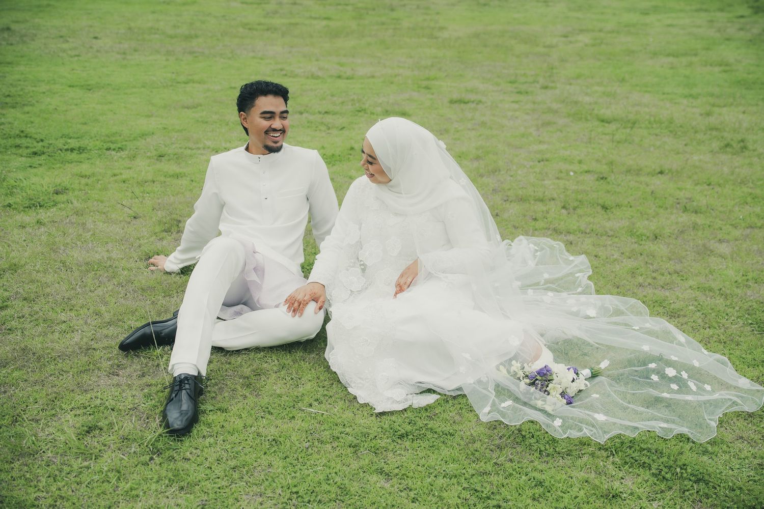 Muslim bride and groom in white attire sitting on green grass, holding hands during outdoor wedding photoshoot.