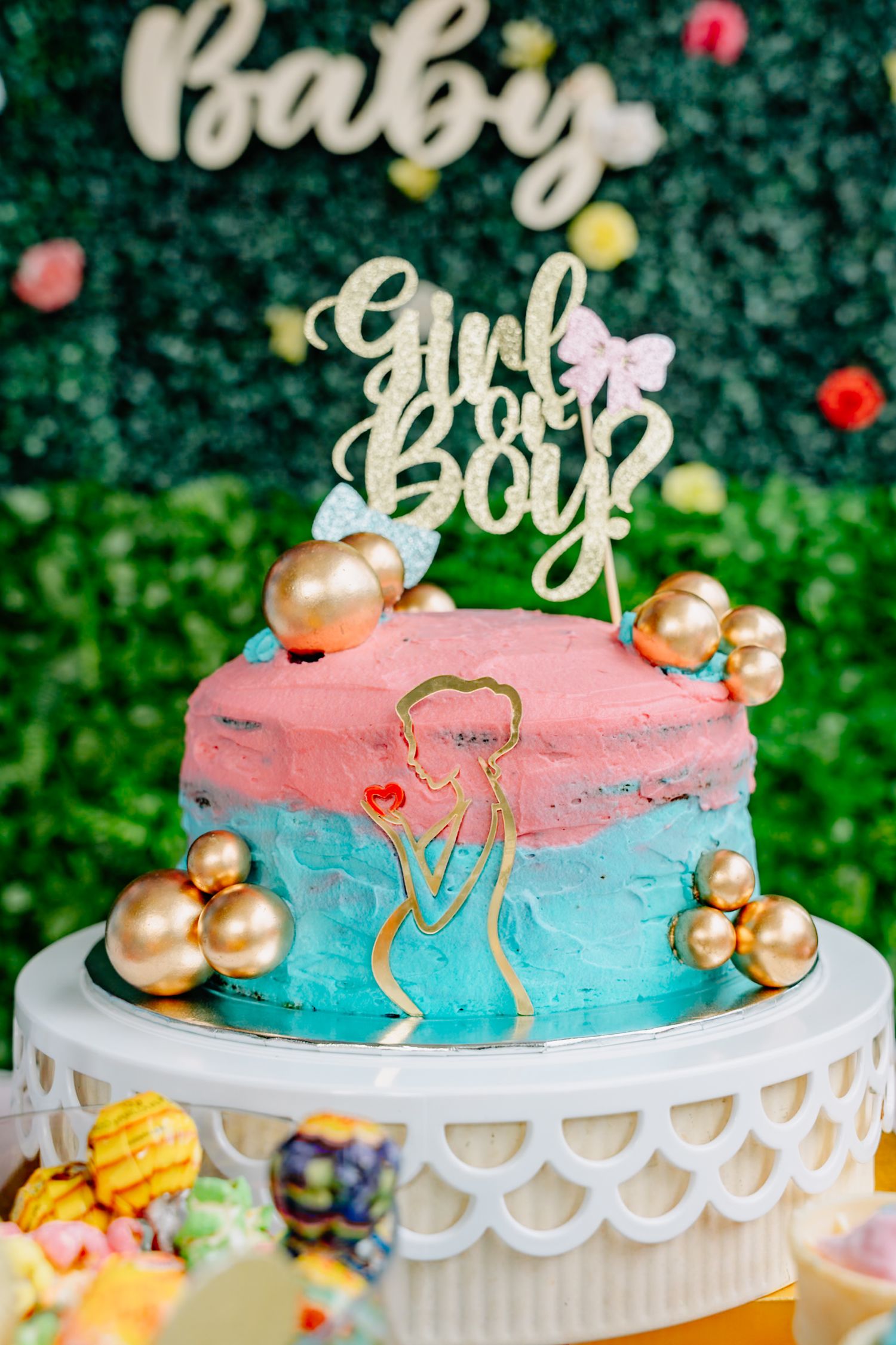 A gender reveal cake decorated in pink and blue ombre frosting with gold accents and pearls on a white cake stand.