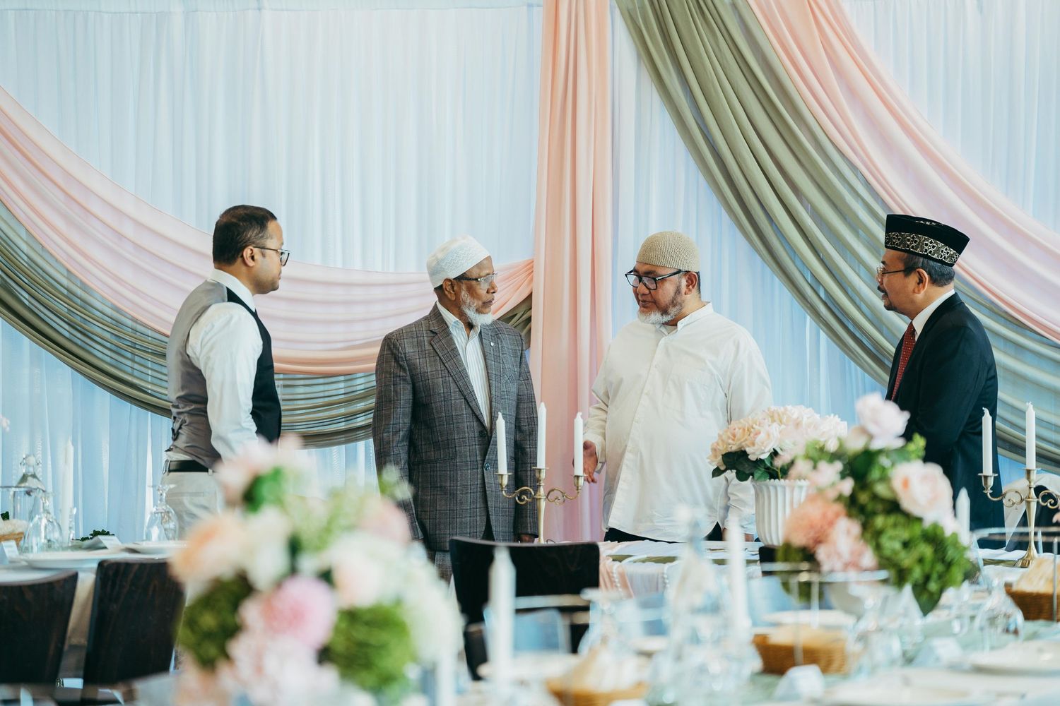 Four men in formal attire converse at an elegantly decorated wedding reception venue with floral centerpieces.