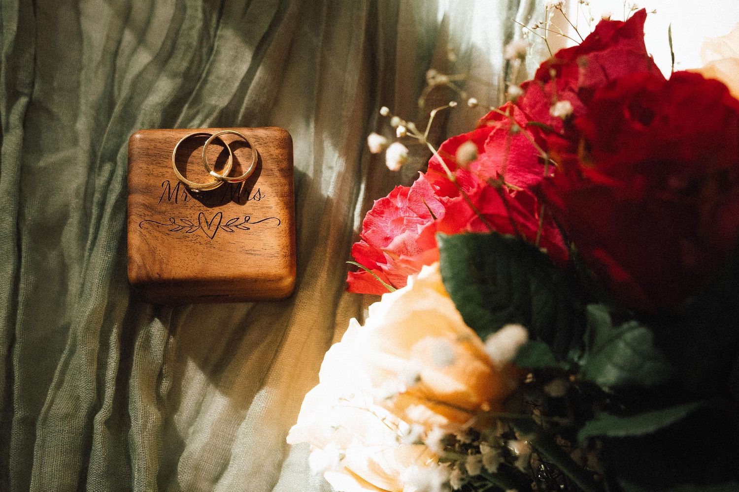 Two gold wedding rings rest on a wooden engraved box beside a vibrant red and white bridal bouquet.