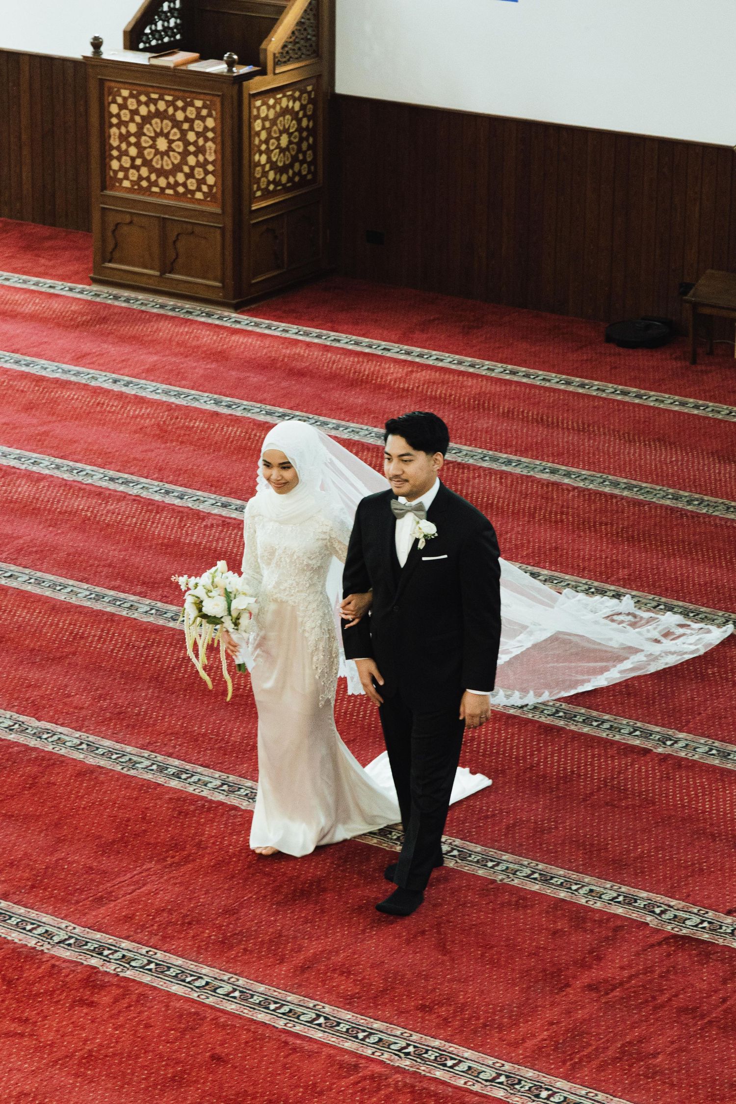 Muslim bride and groom walking down red carpet aisle in mosque after Islamic wedding ceremony.
