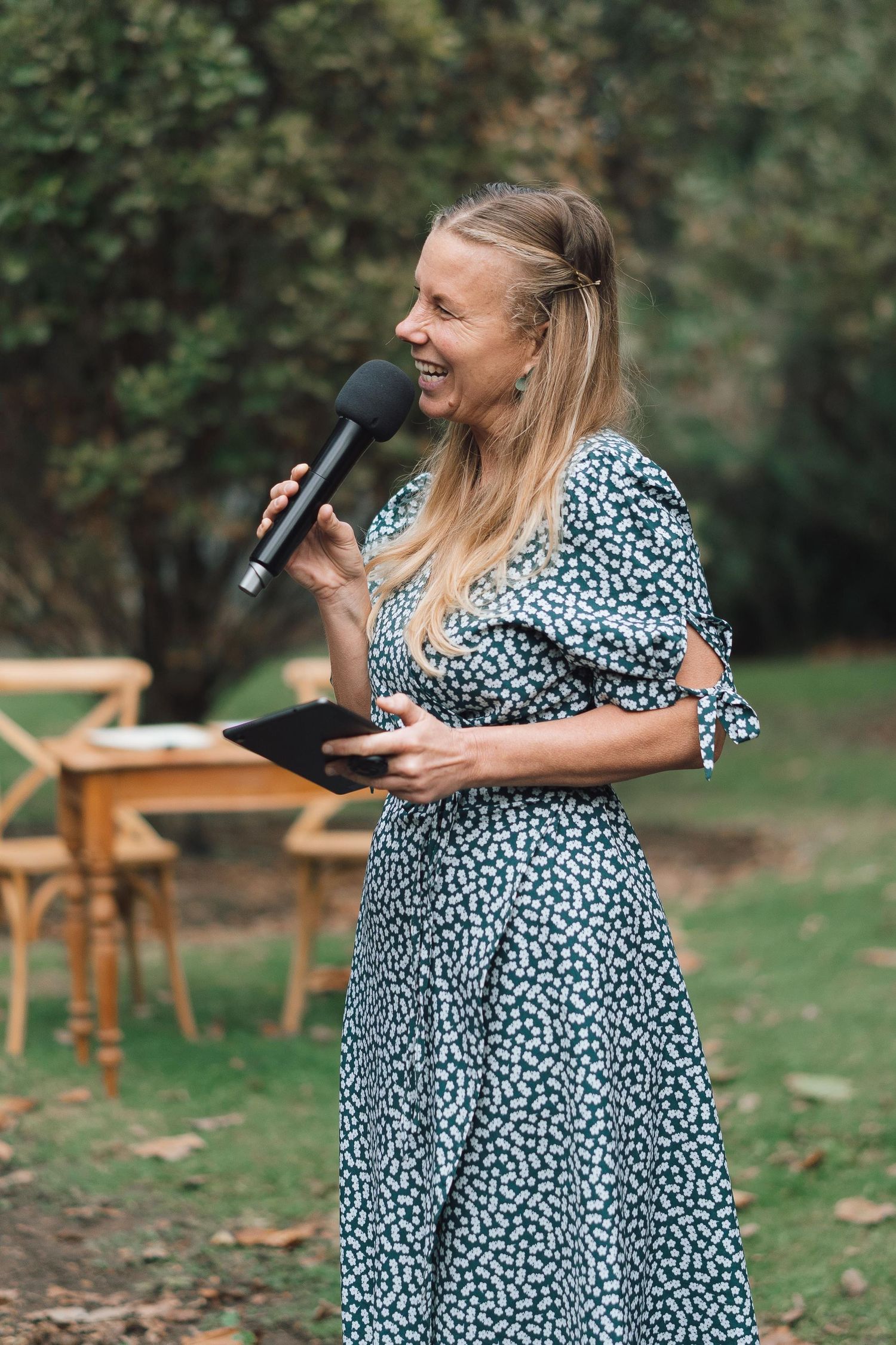 Blonde female wedding celebrant laughing while speaking into microphone at outdoor garden ceremony.