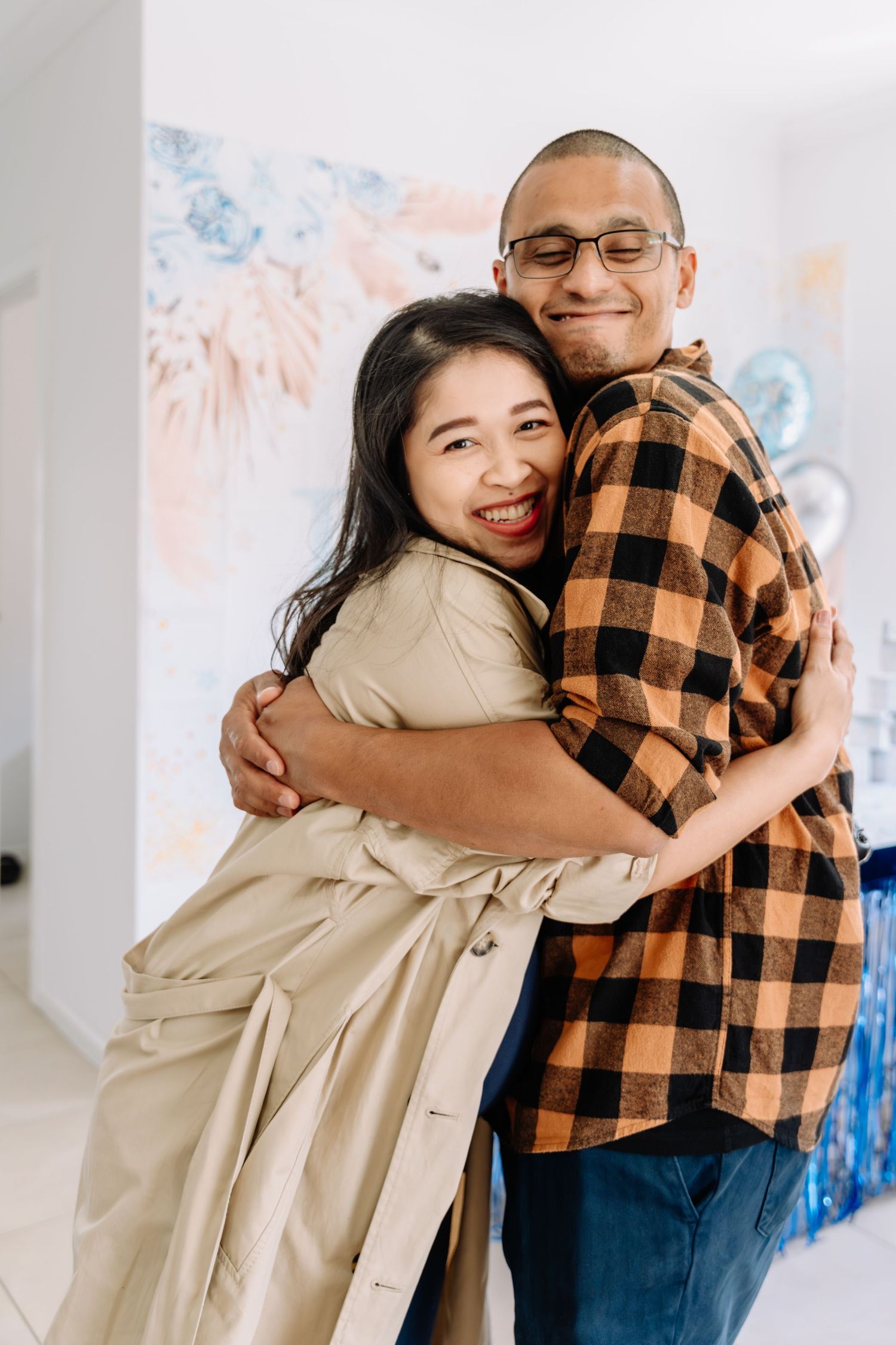 A couple embraces and smiles in front of a decorated wall with blue and pink party decor.