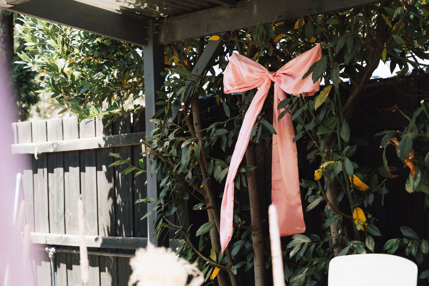 Pink satin bows tied to outdoor patio posts create an elegant garden party decoration against green foliage.