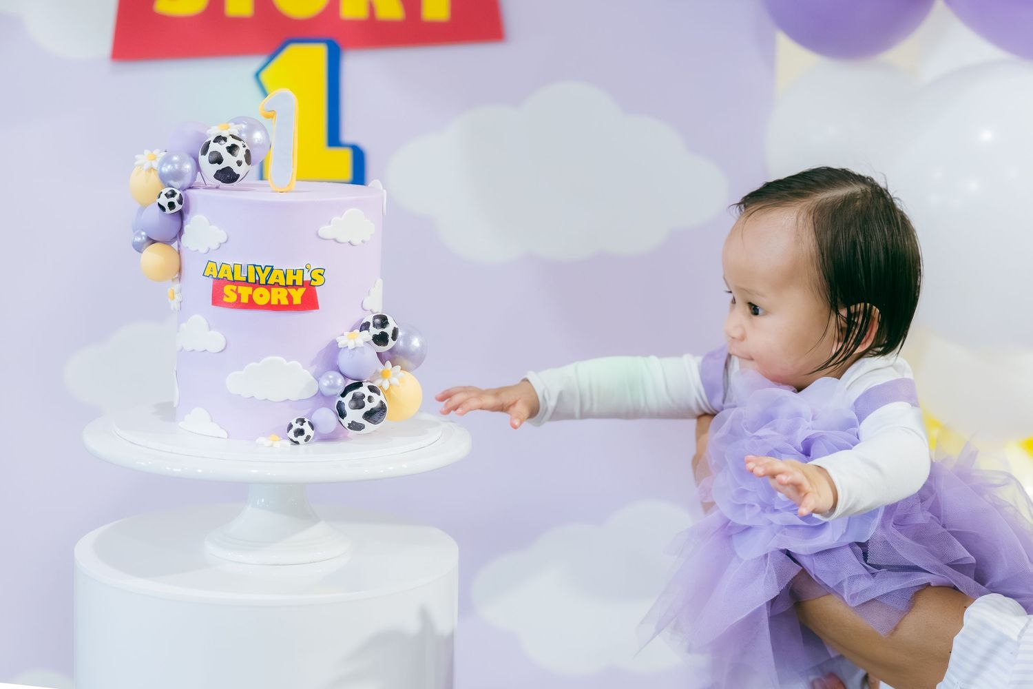 A small child in purple tutu dress reaching toward a white tiered birthday cake with toy story themed decorations.