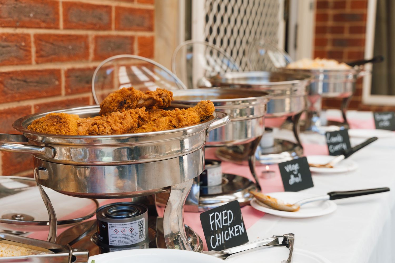 A buffet line of hot food in silver chafing dishes sits on a white tablecloth with serving utensils.