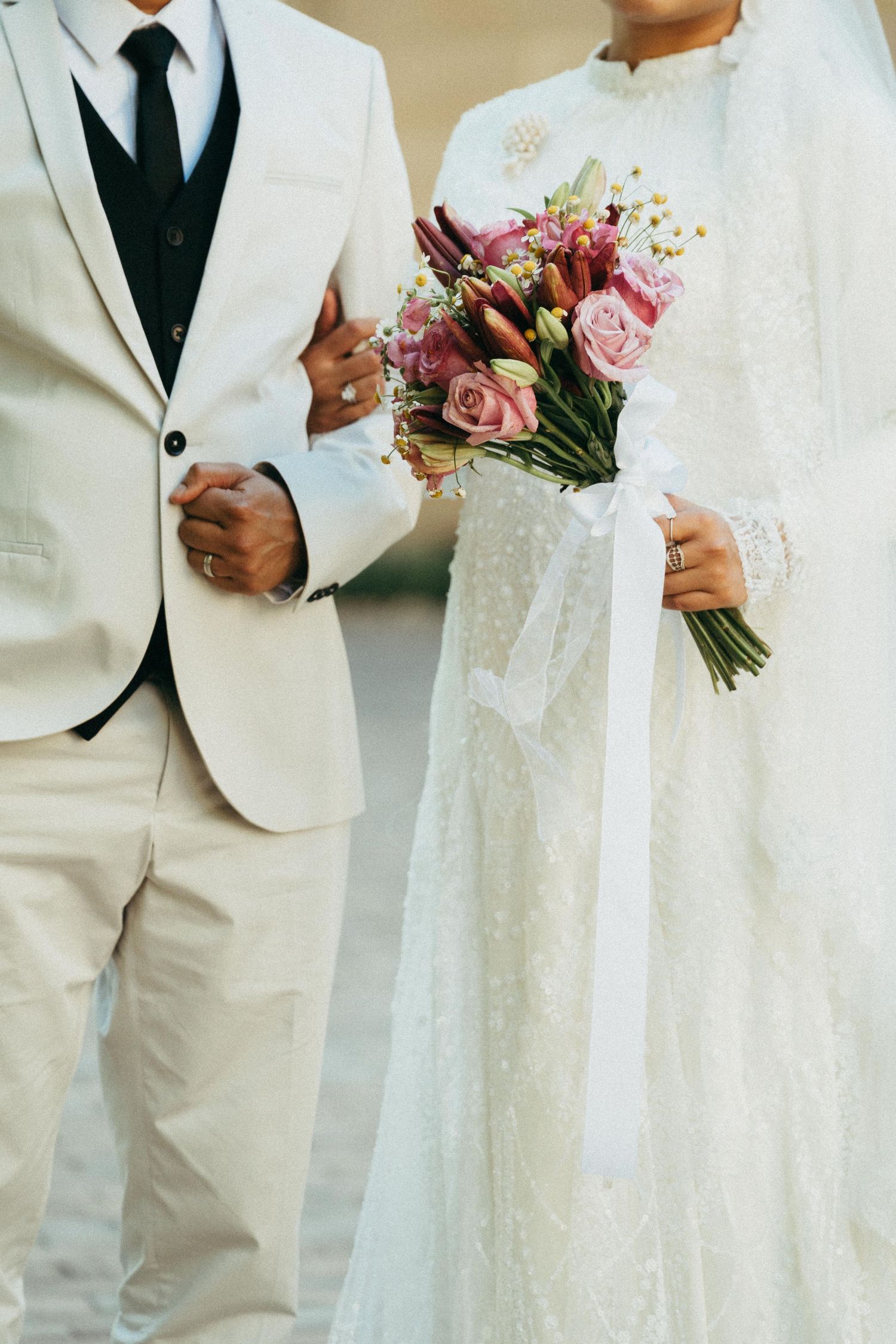 Bride in white lace gown holding colorful bouquet stands beside groom in cream suit at wedding ceremony.