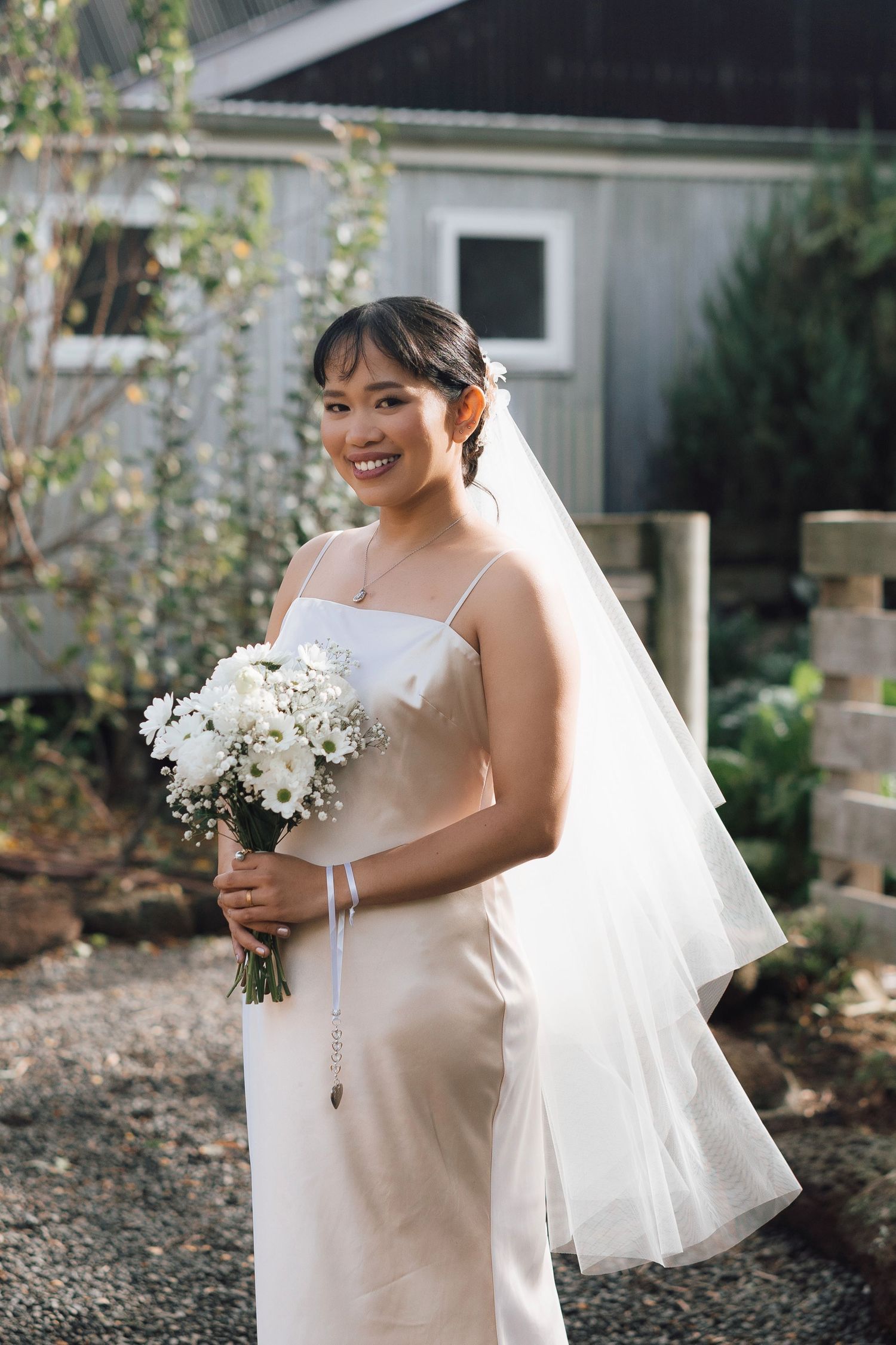 Smiling bride in a satin slip dress and veil holding a white floral bouquet outdoors.