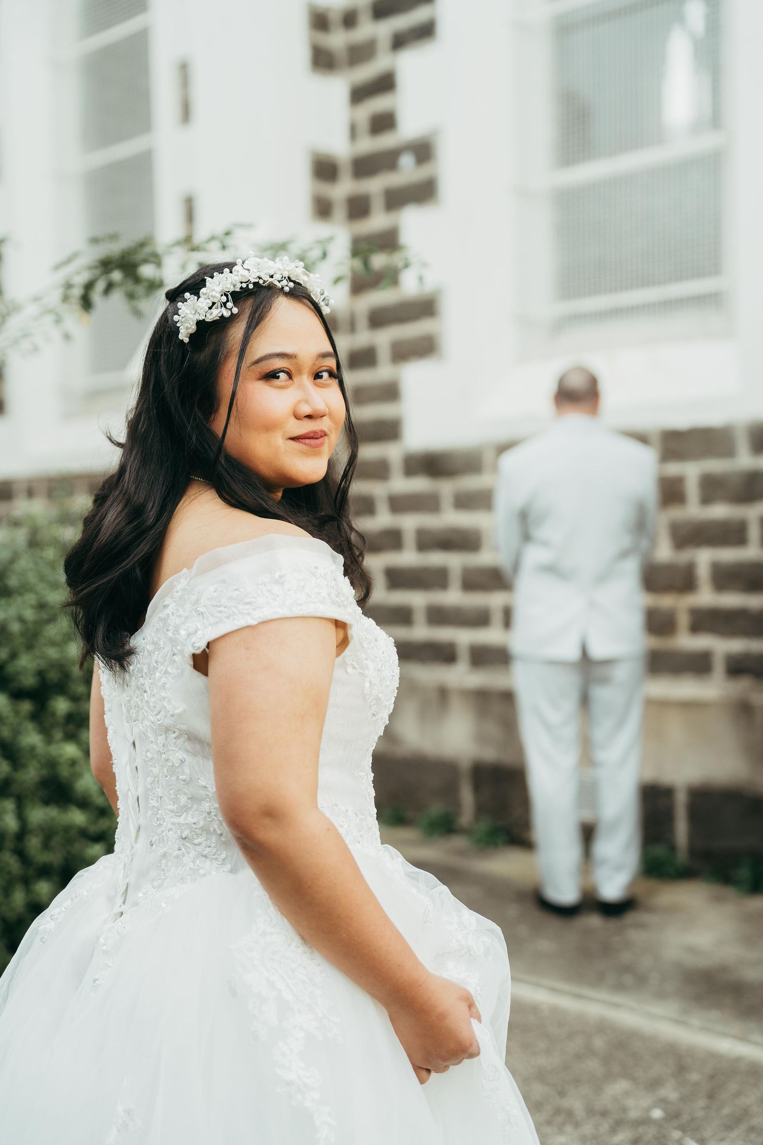 Bride in white lace off-shoulder gown and floral crown smiles outdoors, groom in light suit stands facing away.