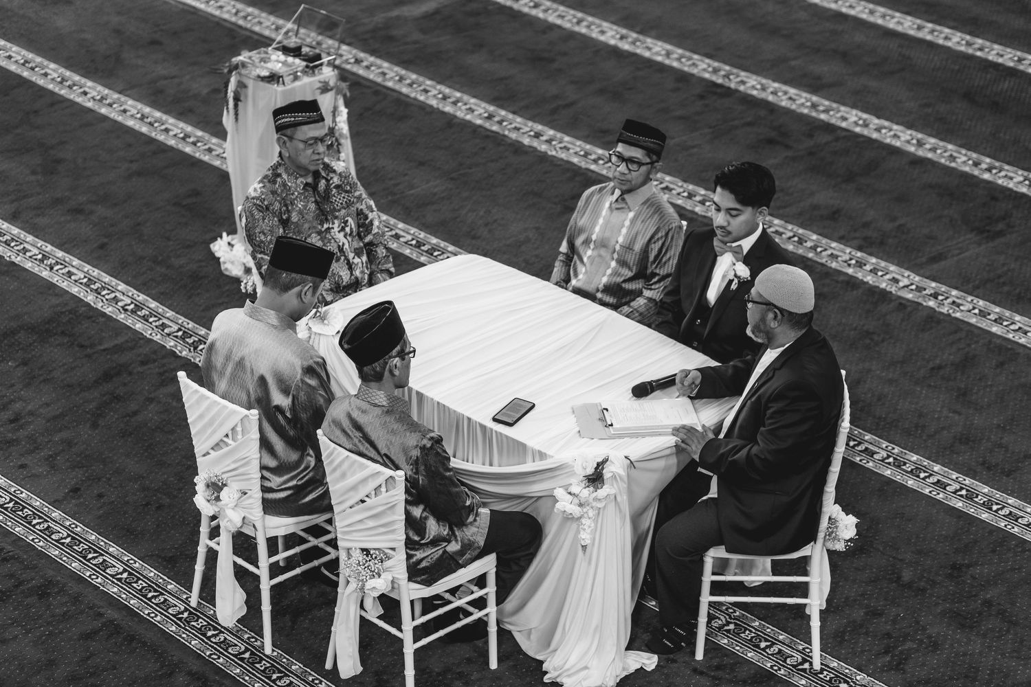 Black and white photo of a traditional Islamic wedding ceremony with men seated at a white table on a patterned mosque floor.