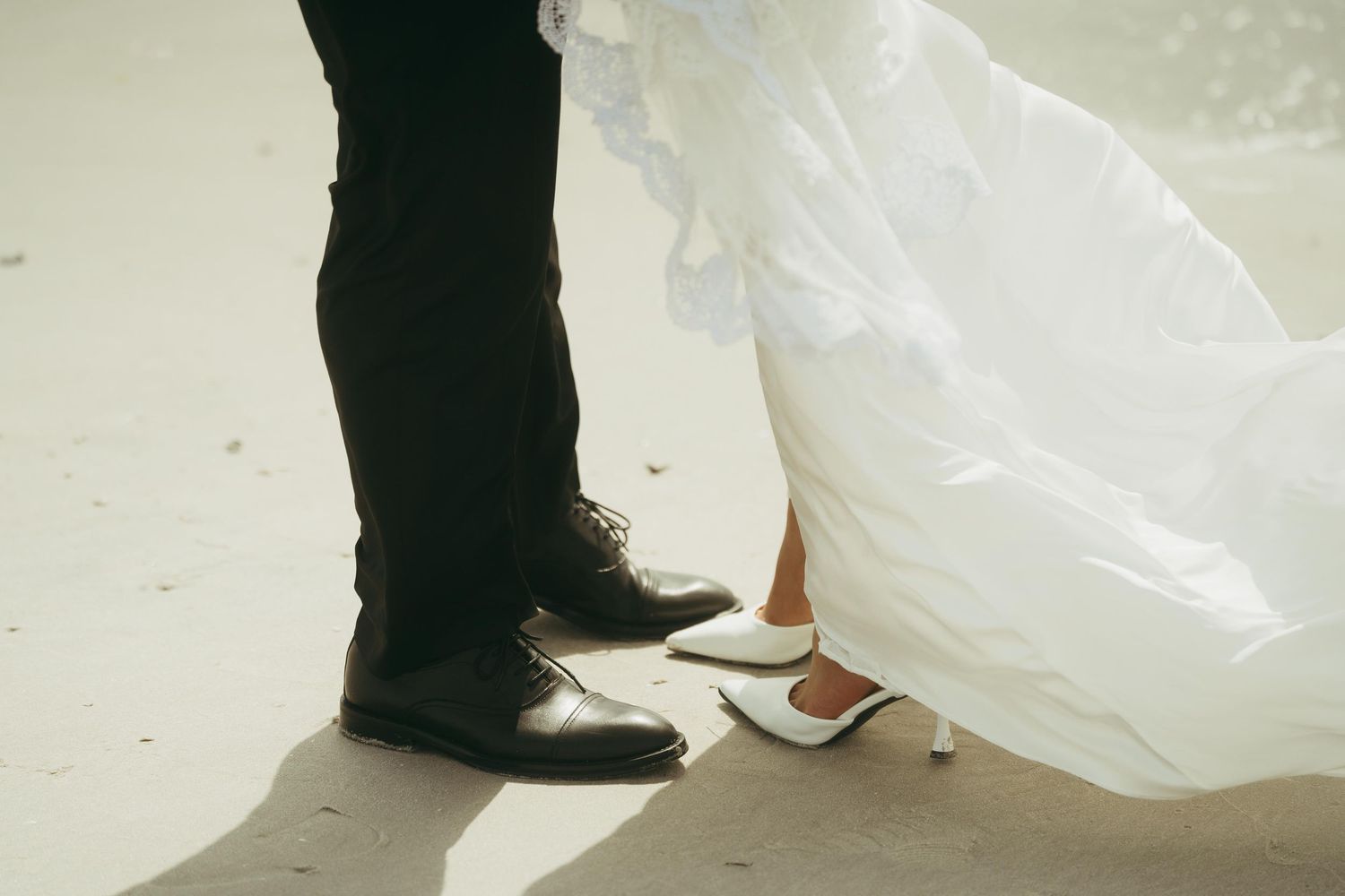 Close-up of bride and groom feet in wedding shoes standing on sandy beach.