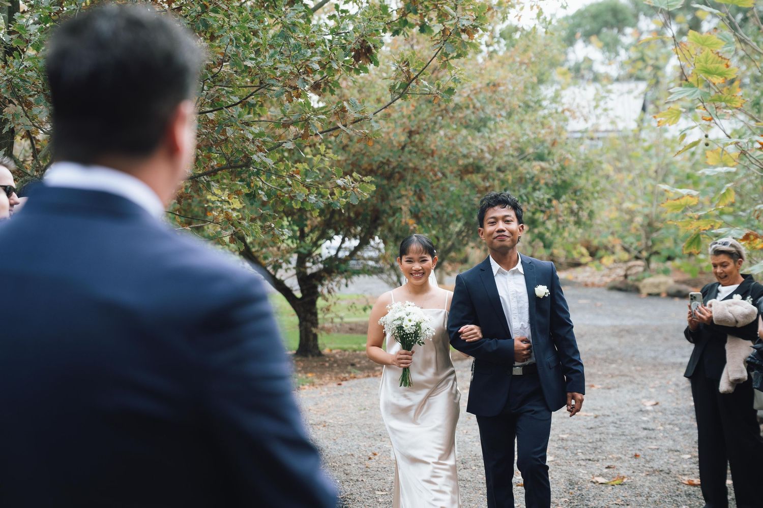 Bride in white gown walks with groom toward guest at elegant outdoor wedding ceremony.