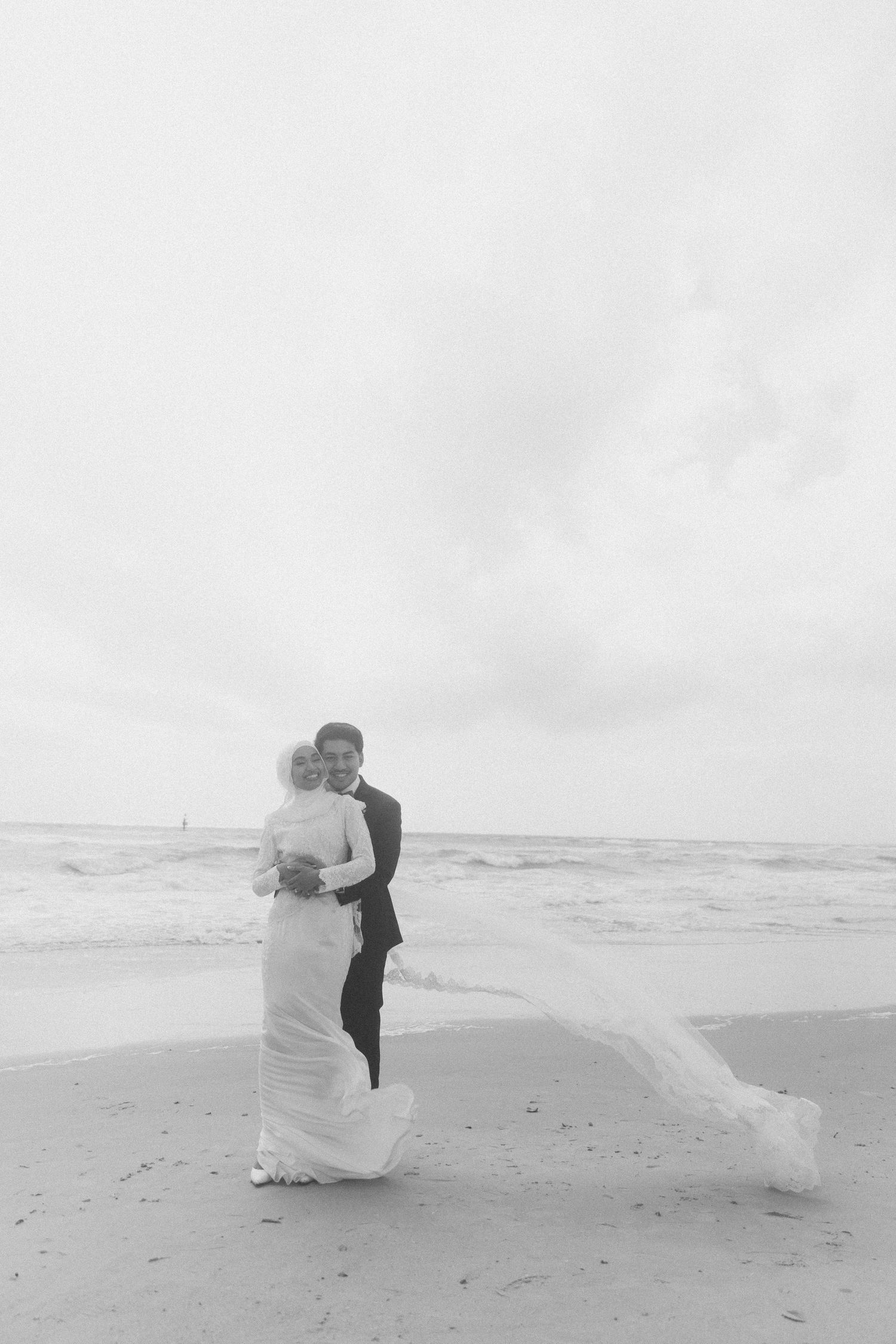A bride and groom embrace on a beach in a black and white photo with waves in the background.