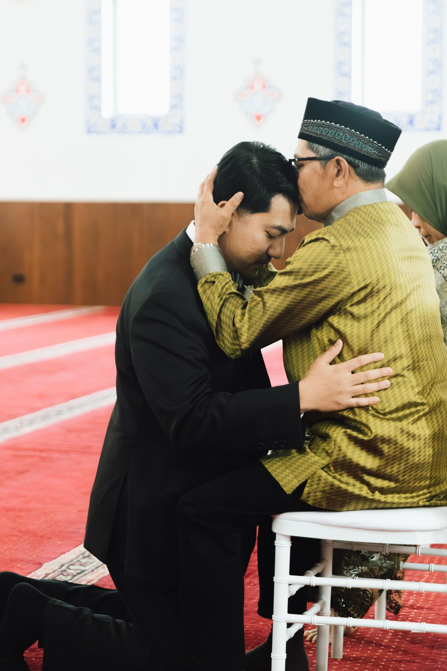 A young man kneels to receive a blessing from an elder in traditional attire inside a mosque.