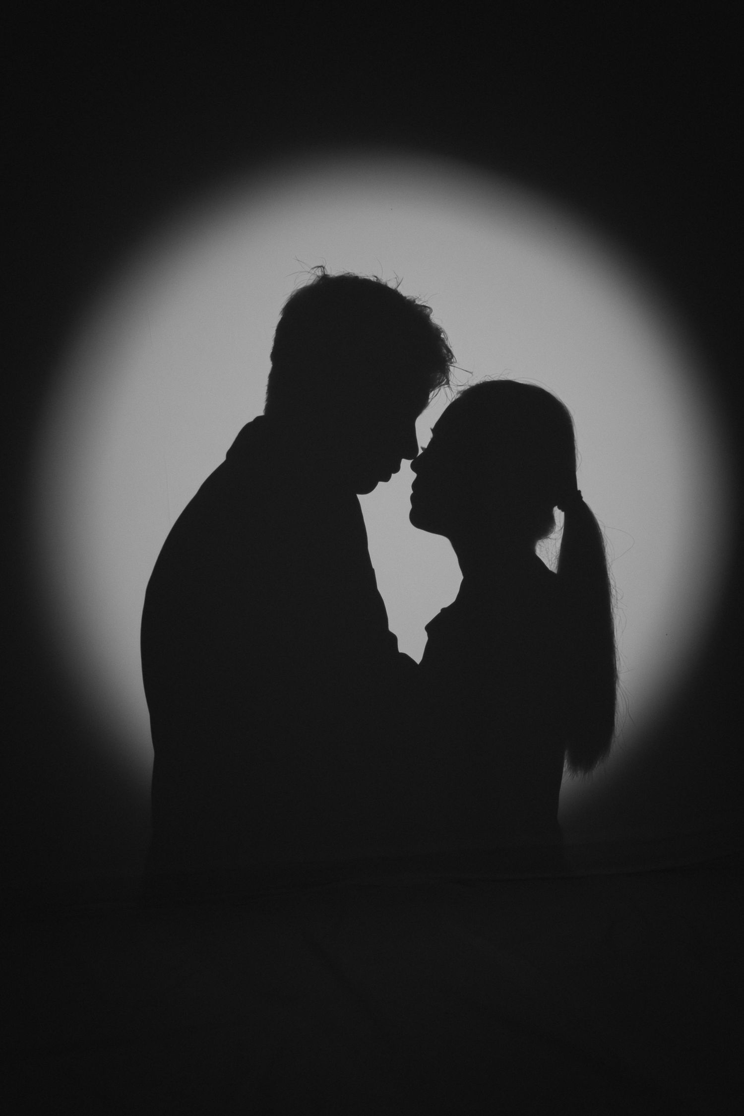 Romantic silhouette of a couple facing each other against a glowing circular light in black and white.