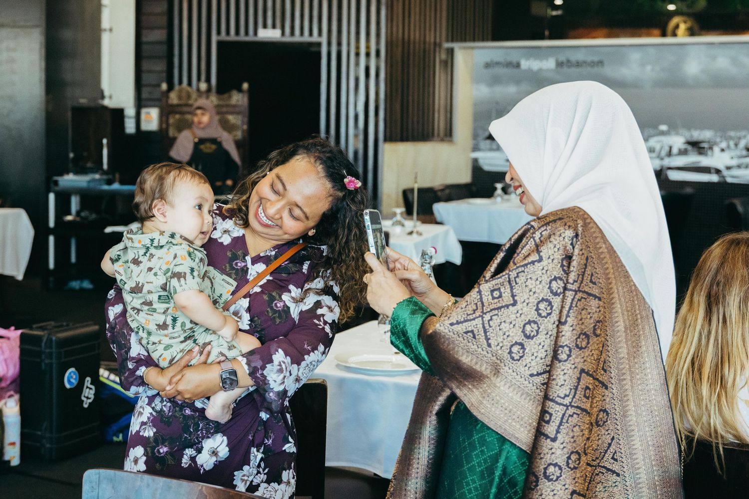 Woman in floral dress holding baby smiles as guest in hijab photographs them at wedding reception.