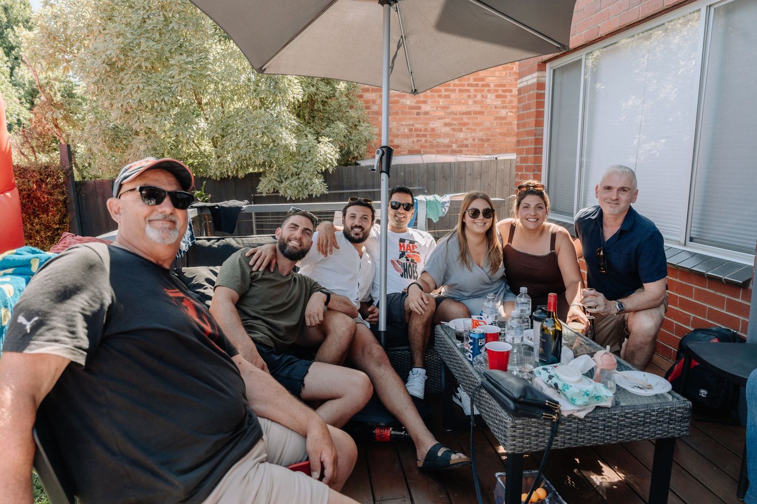 Outdoor patio gathering with people seated under umbrellas at tables during a social event.