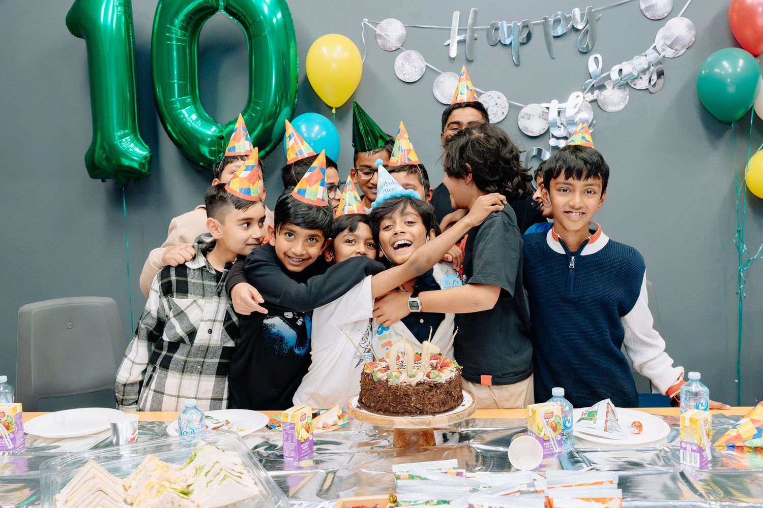 Friends gather around a birthday cake at a celebration with green and yellow balloons in the background.