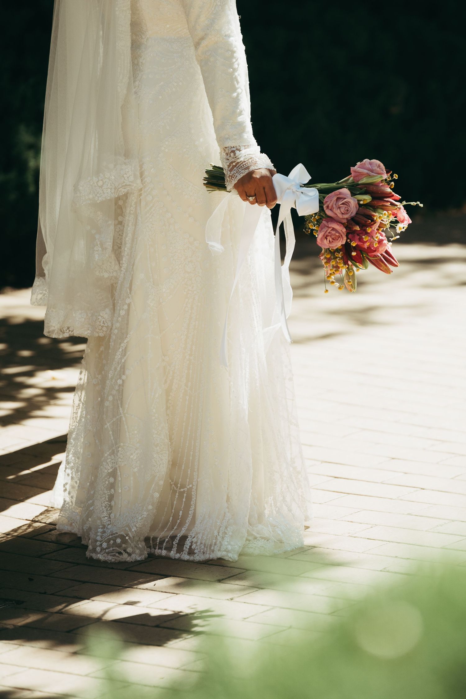 Close-up details of a flowing white lace wedding dress with delicate floral bouquet in soft natural lighting.