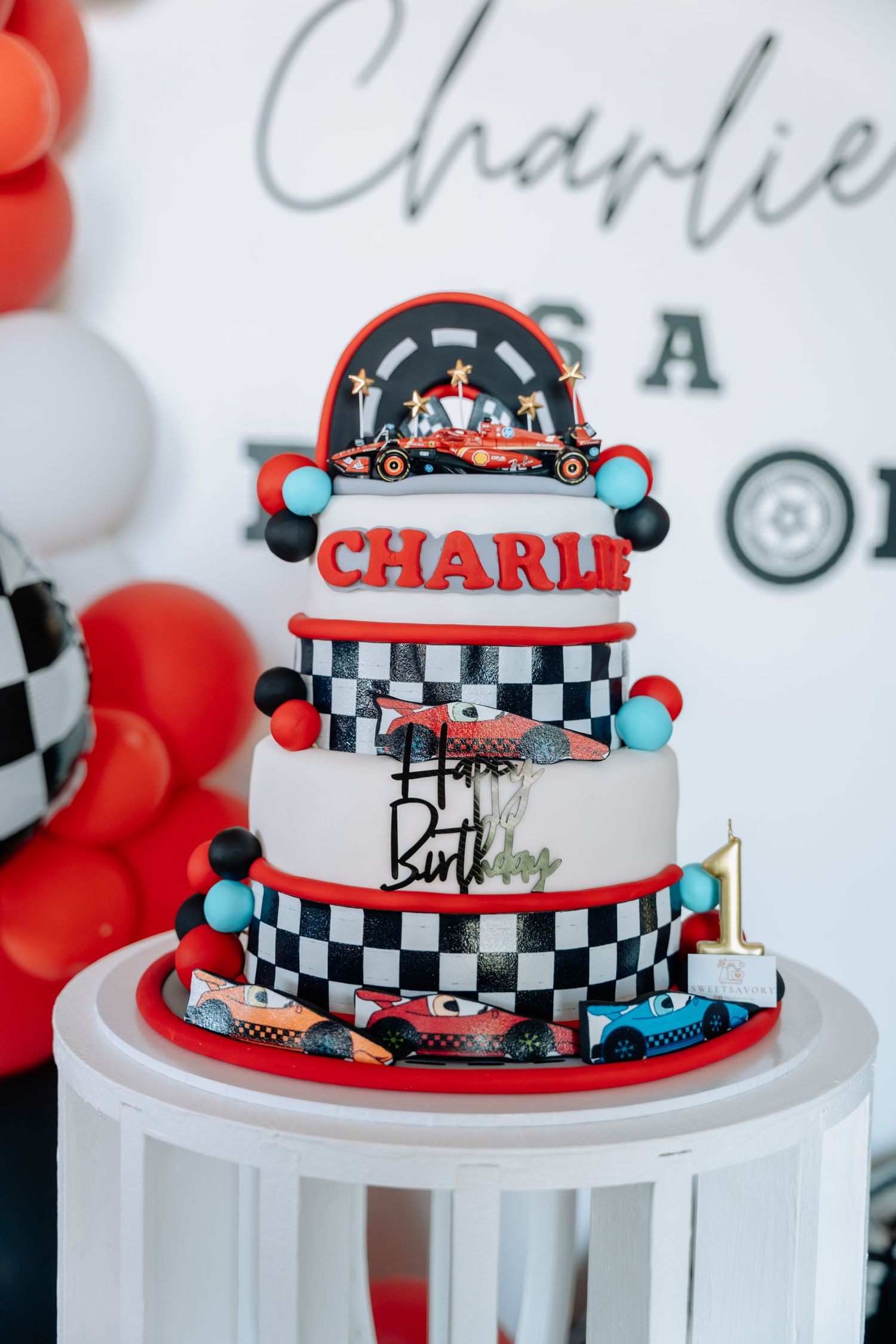 Racing themed birthday cake with red and checkered pattern tiers on a white pedestal stand.