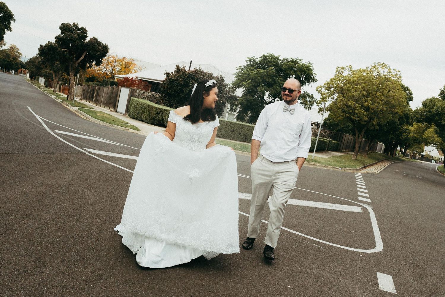 Bride in white gown and groom in white shirt walk together on a suburban street on their wedding day.