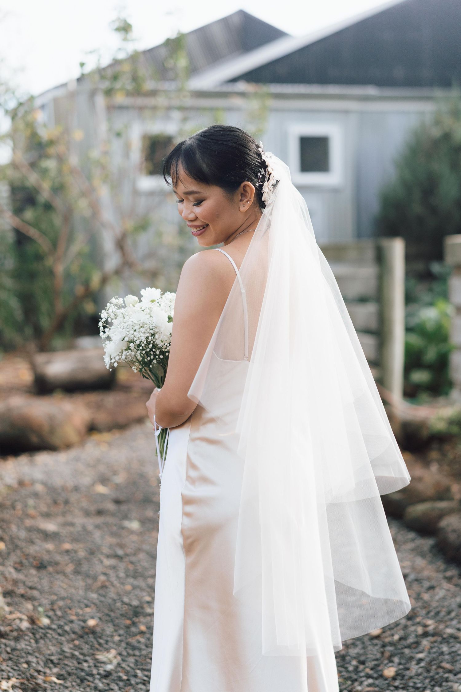 Smiling bride in white satin gown and veil holds baby's breath bouquet outdoors.