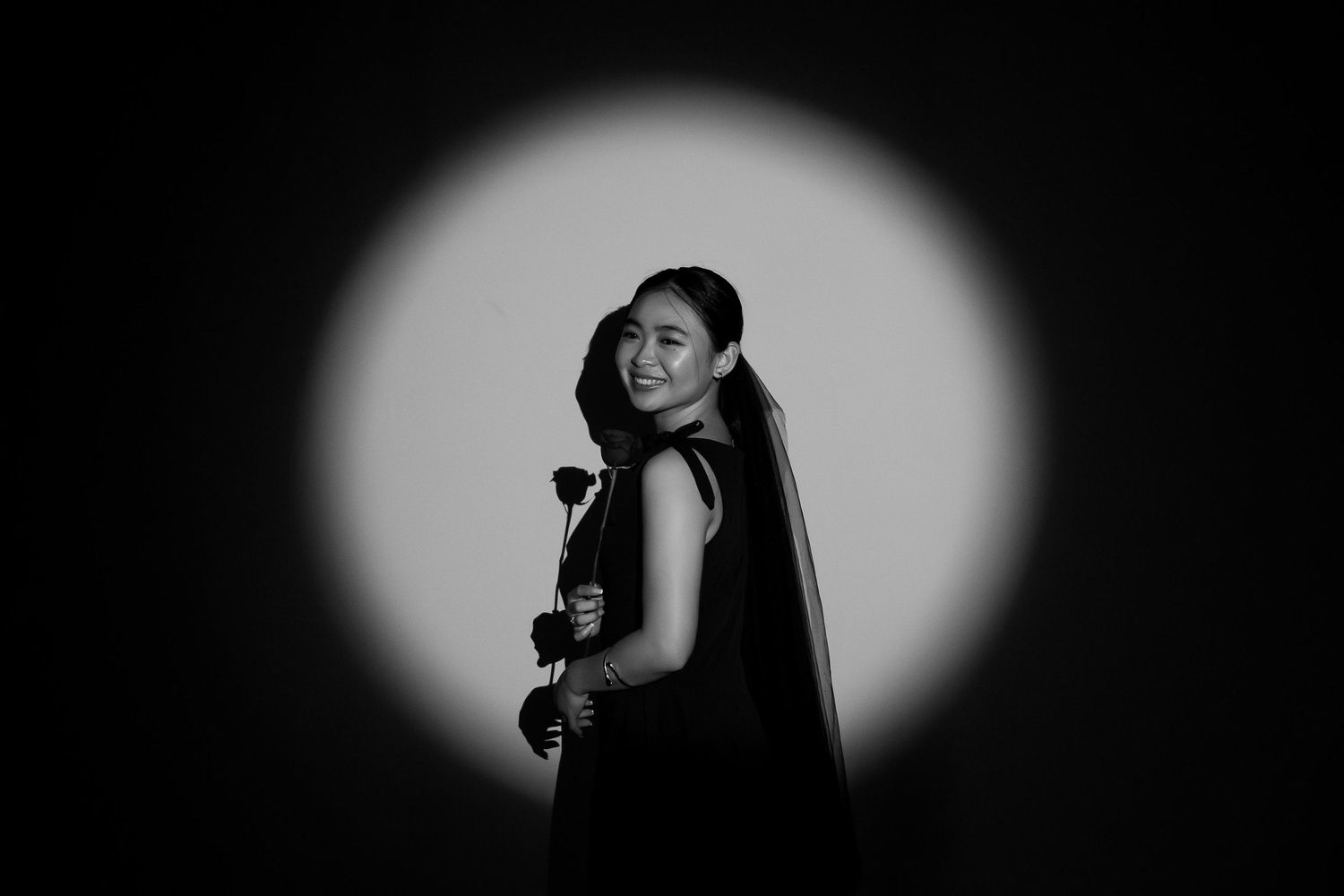 Woman in black dress holding a rose, smiling in a dramatic circular spotlight, black and white.