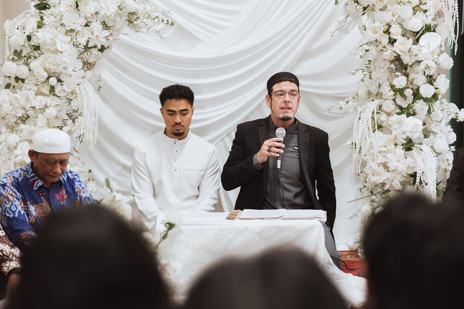 Three men at a wedding ceremony table with white floral backdrop, one speaking into microphone.