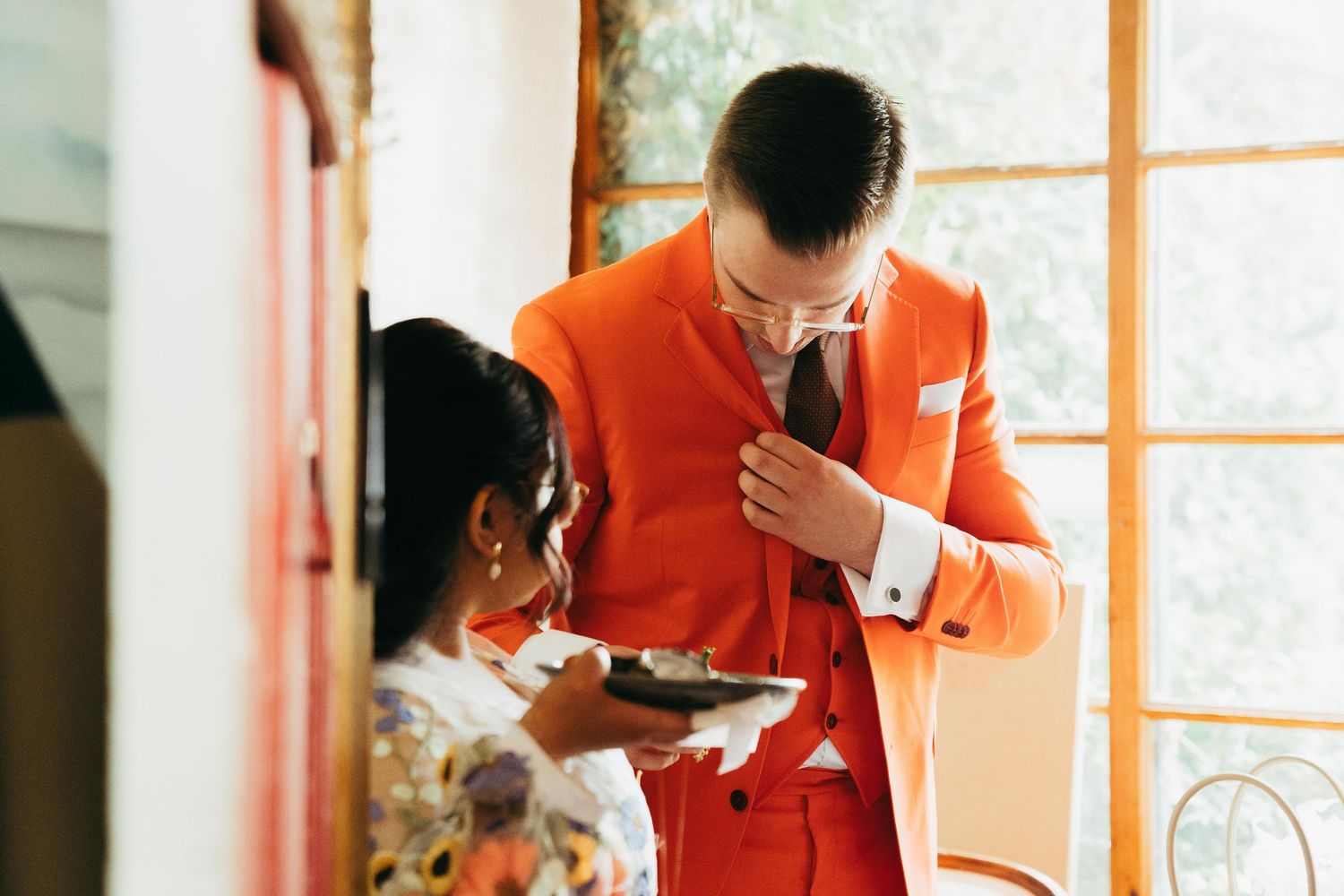 Groom in bold orange suit adjusting tie before wedding ceremony indoors.