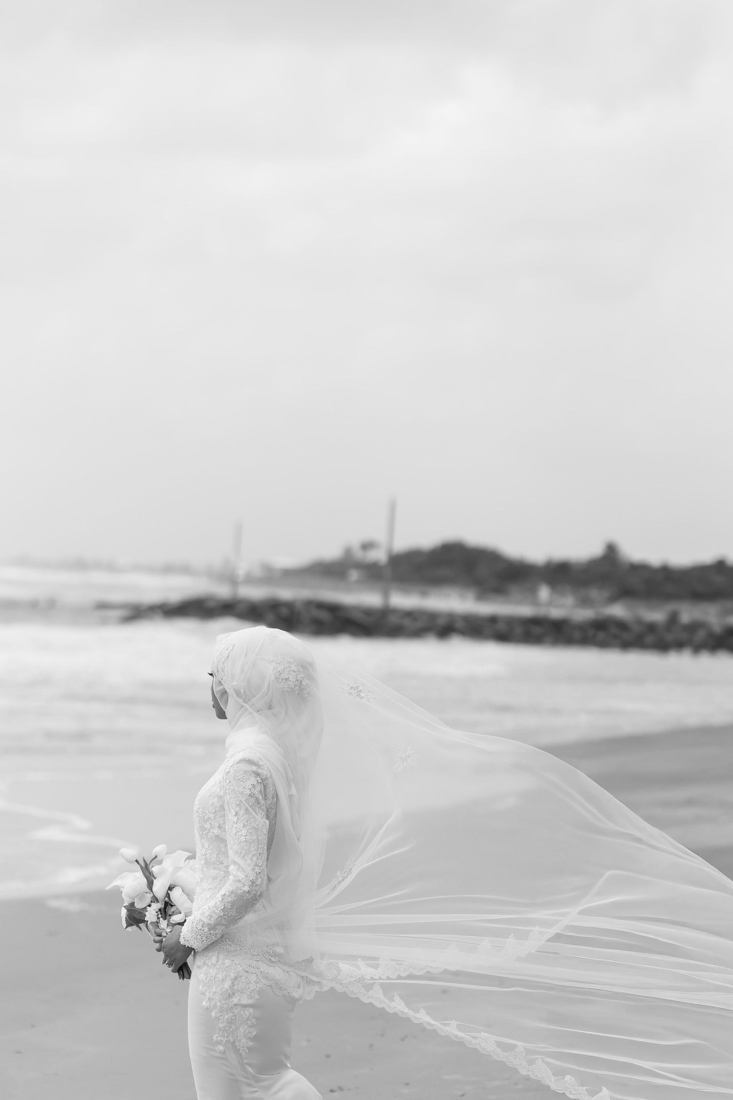 Black and white photo of Muslim bride in white gown with flowing veil on beach.