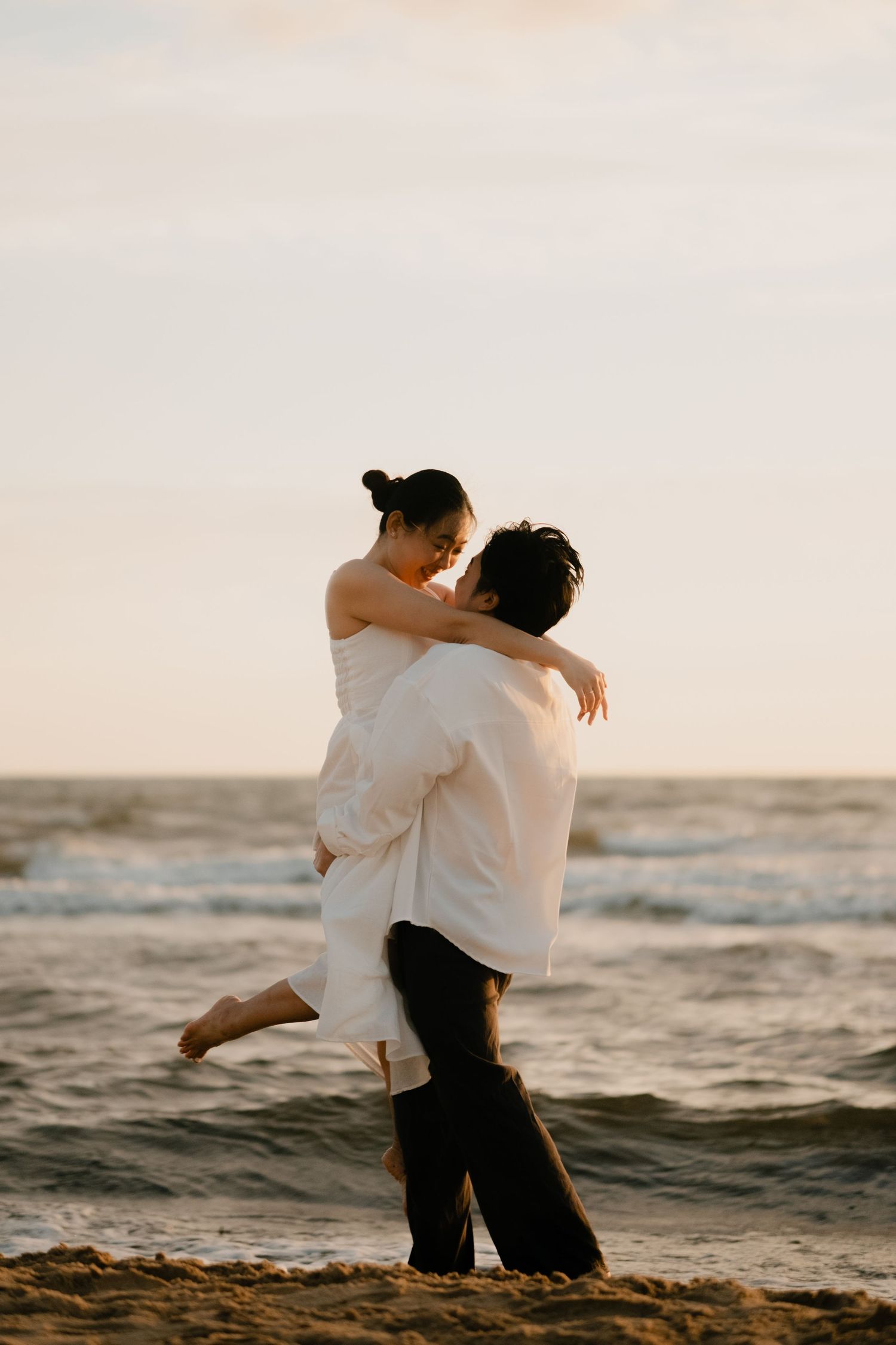 Romantic couple embracing on a beach at sunset, man lifting woman in white dress by the ocean.