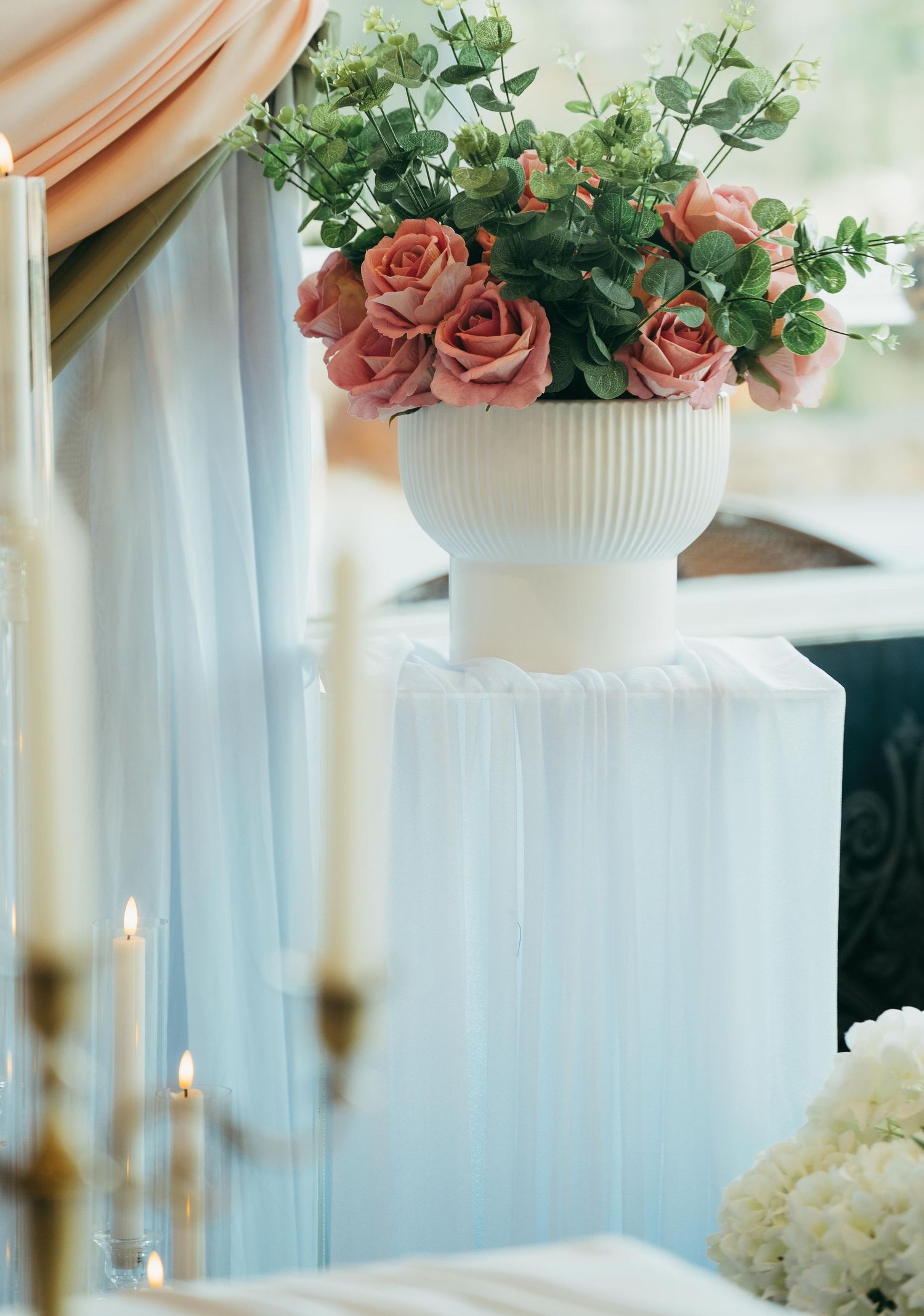 Pink roses and eucalyptus in white ribbed vase on draped pedestal with candles at a romantic wedding venue.