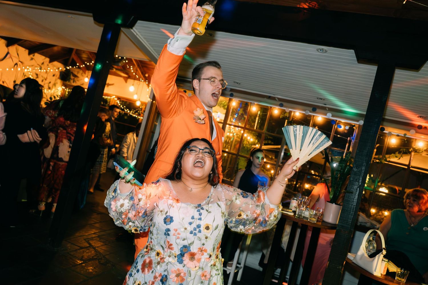 Couple celebrating at outdoor wedding reception, man in orange suit and woman in floral dress.