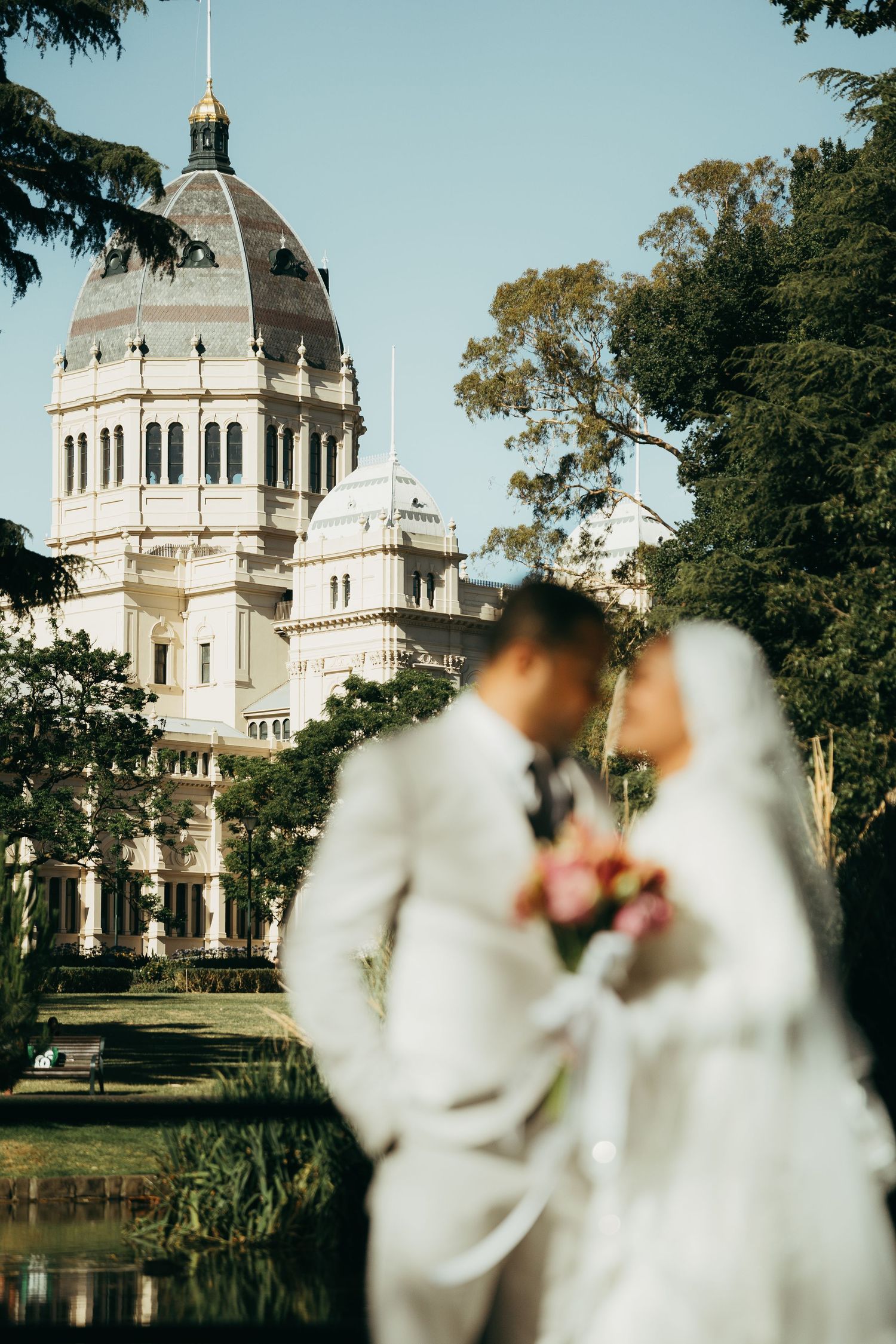 A romantic wedding couple in white stands before the majestic Royal Exhibition Building in Melbourne on a sunny day.