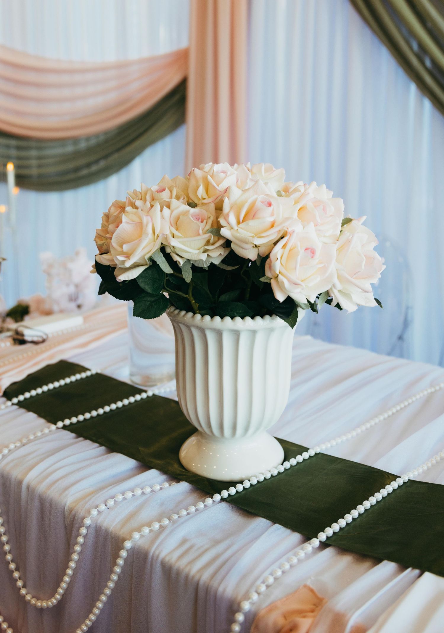 Elegant white urn vase with cream roses centerpiece on pearl-adorned dark green table runner at a wedding reception.