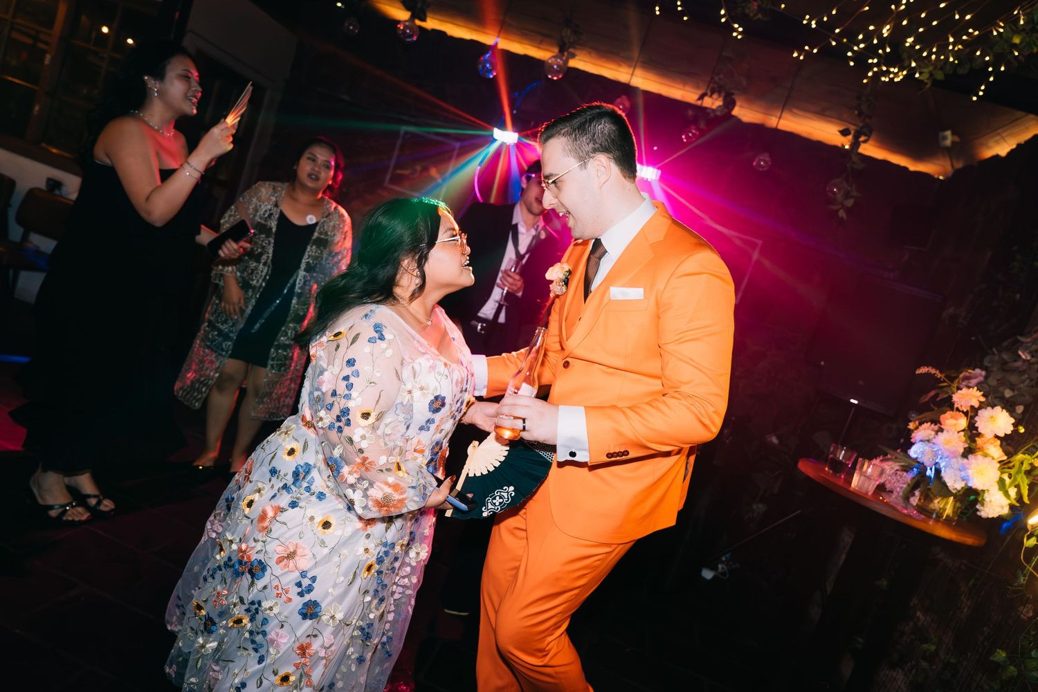 A bride in a floral dress dances with a groom in an orange suit at a wedding reception.