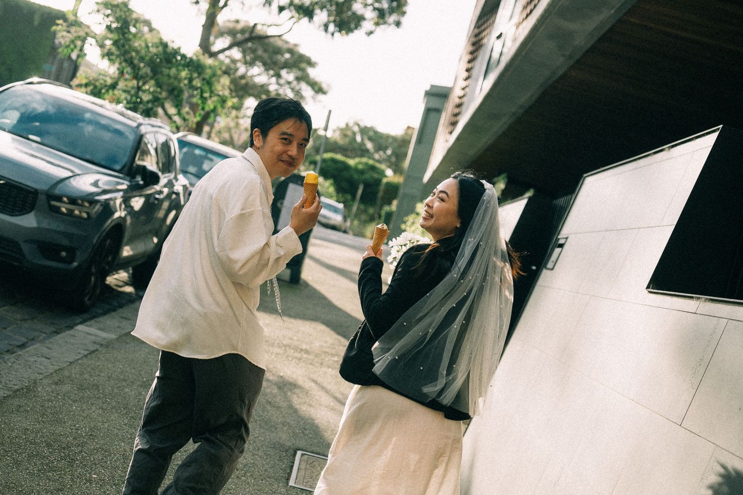 Two people smiling and eating snacks on a city street beside parked cars and a building.