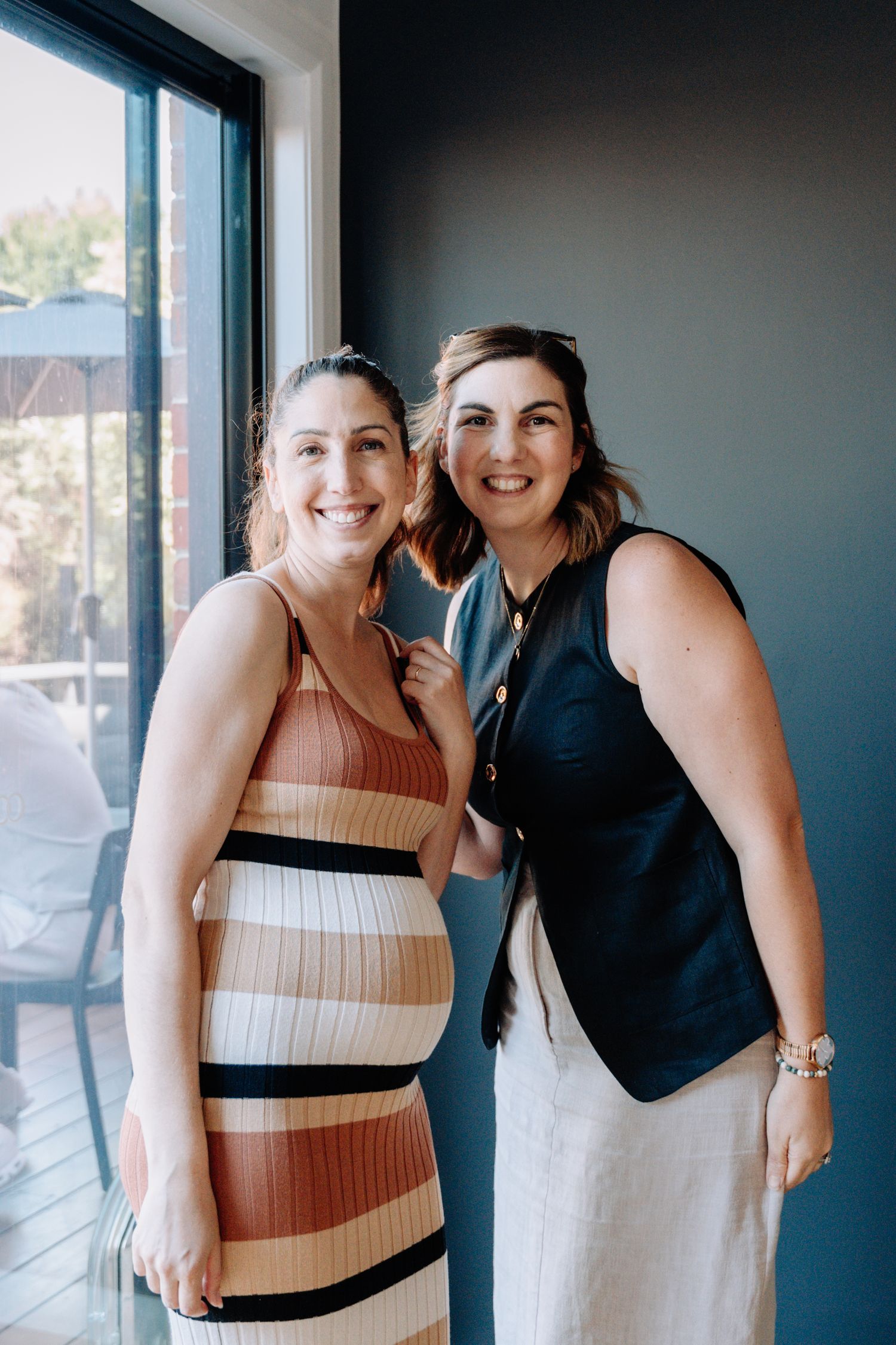Two people in casual attire pose together by a window in an indoor setting with natural lighting.