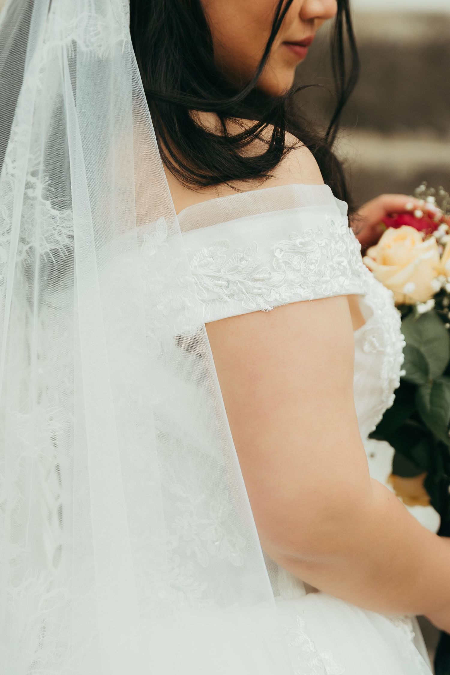 Bride in white off-shoulder lace wedding gown with veil holding a bouquet of roses.