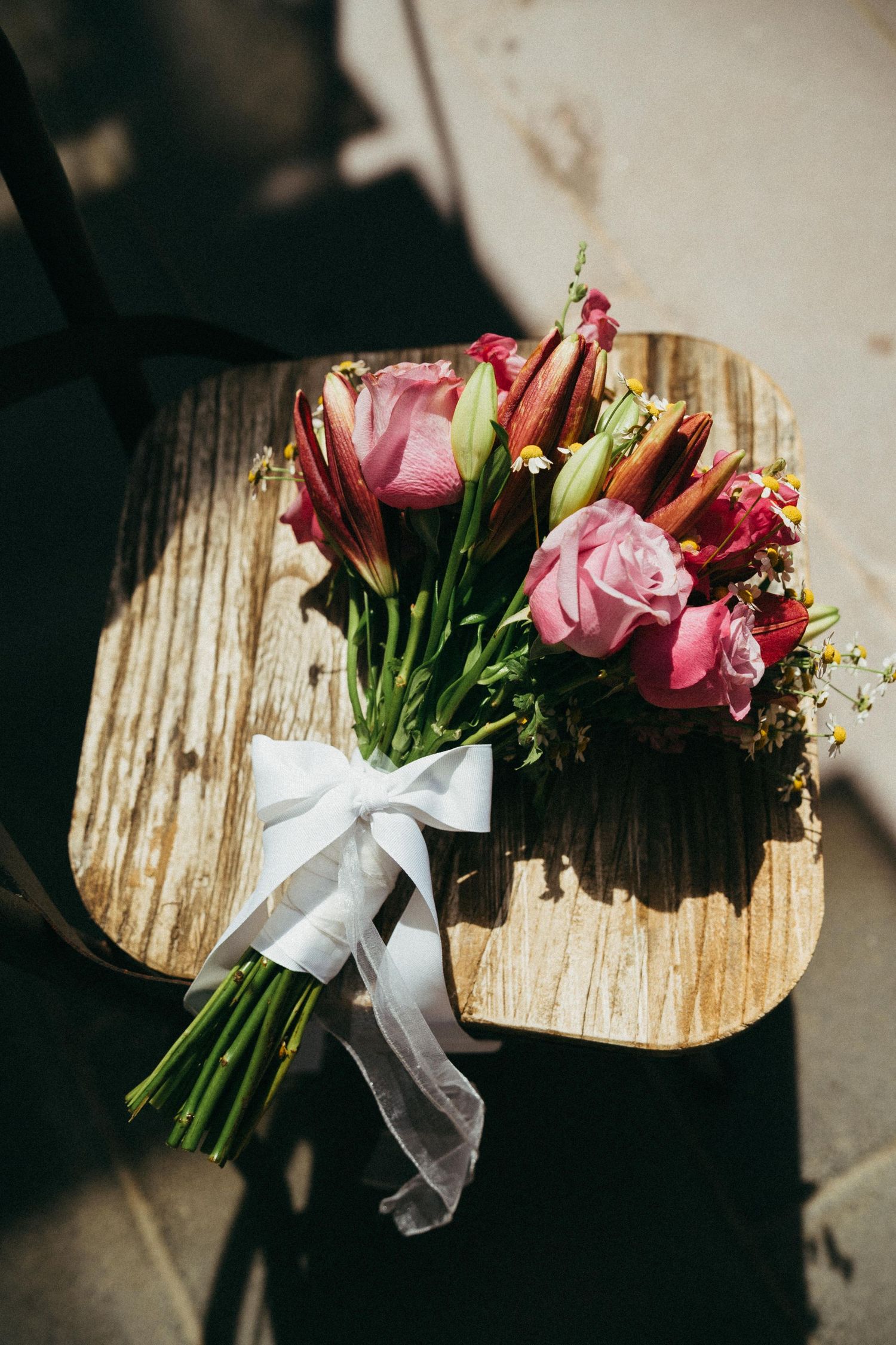 Pink roses and tulips bridal bouquet tied with white ribbon bow resting on rustic wooden chair in sunlight.