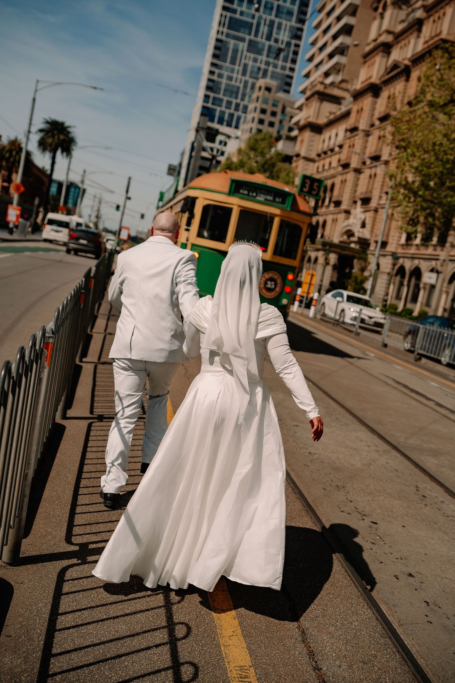 Bride and groom in white walk hand-in-hand on a city street with a green tram in the background.