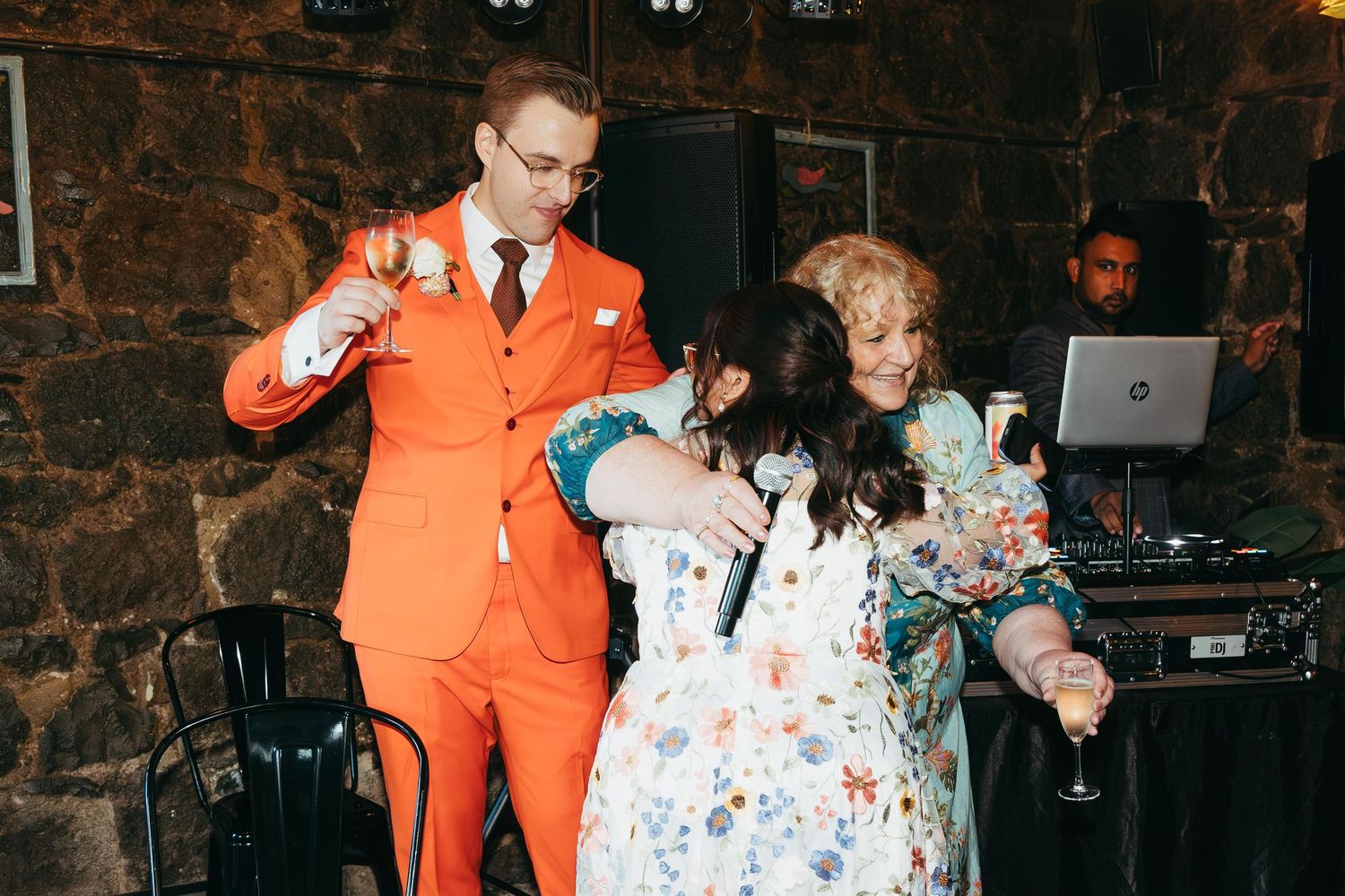Man in orange suit dancing with two women in floral dresses at a wedding reception.