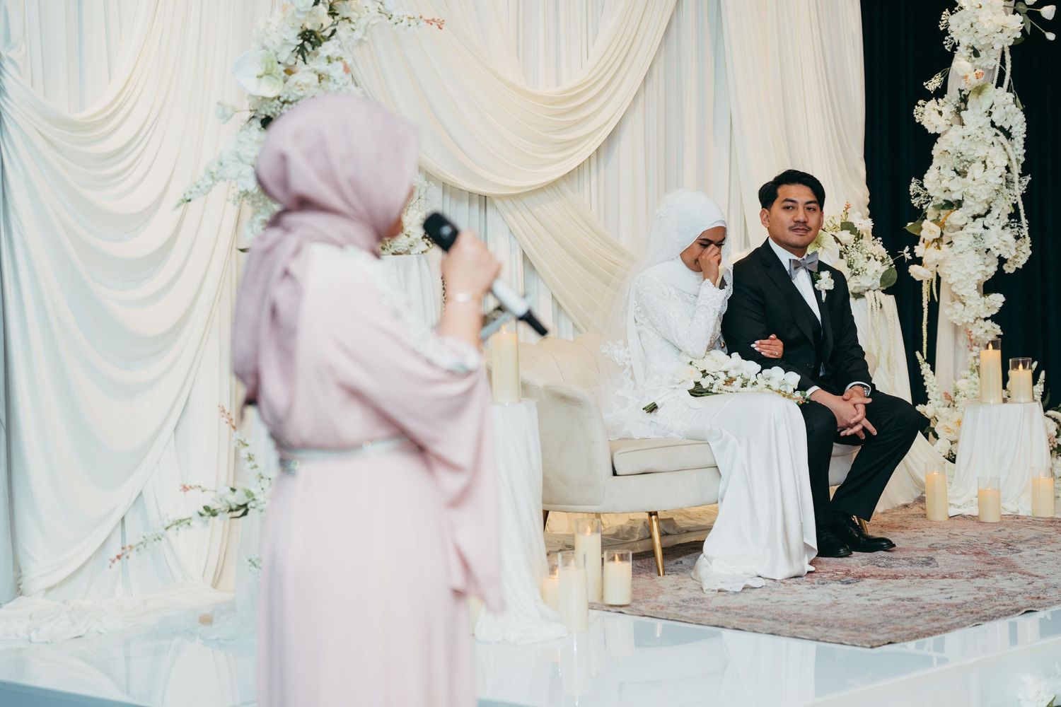 A woman in hijab speaks at a Muslim wedding ceremony while the bride and groom sit on a decorated stage.