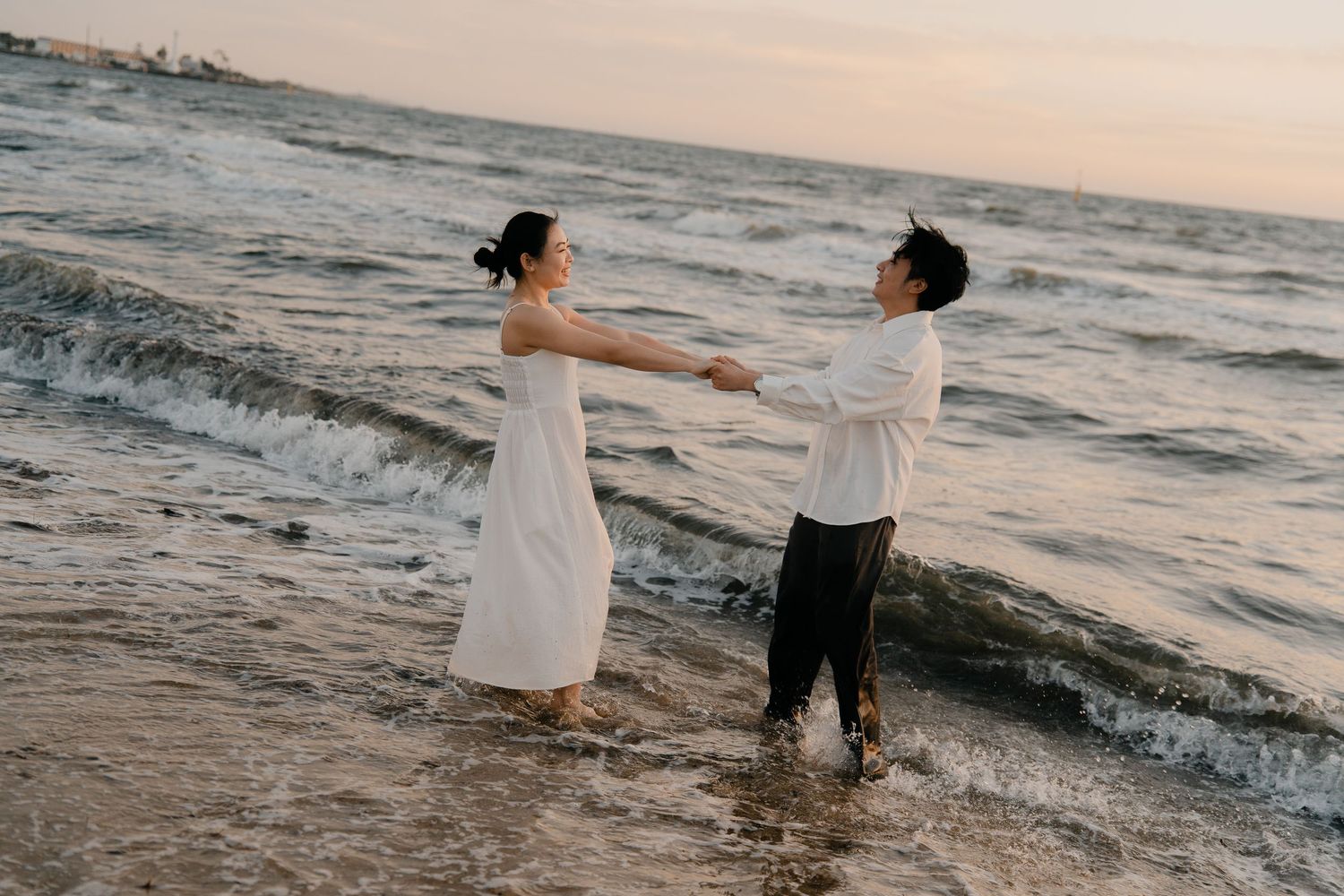 Couple holding hands dancing in ocean waves at sunset beach, woman in white dress, romantic engagement photo.